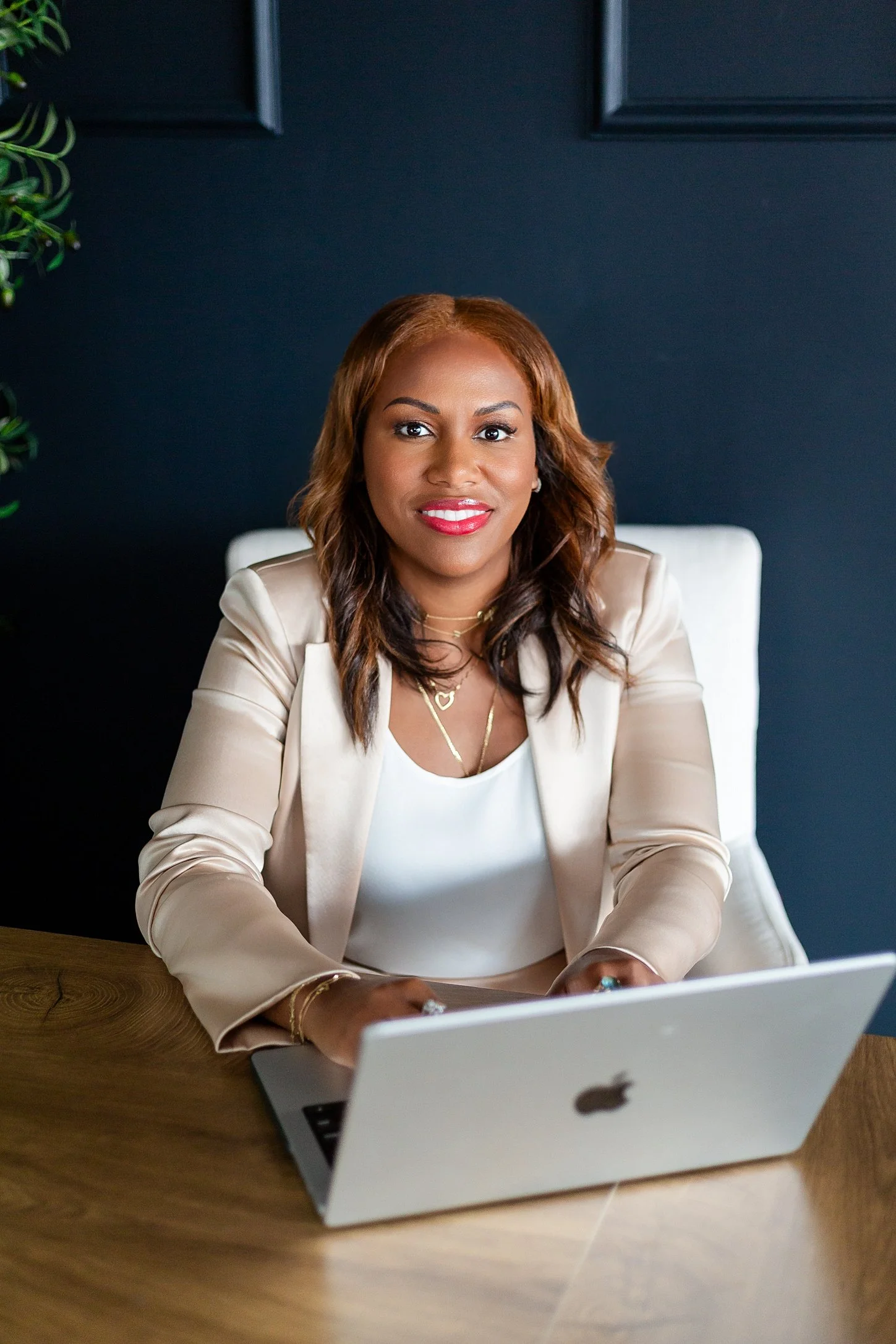A woman with brown hair and red lipstick sitting at a wooden desk with a silver MacBook laptop, wearing a white top and beige blazer, smiling at the camera against a dark blue wall background.