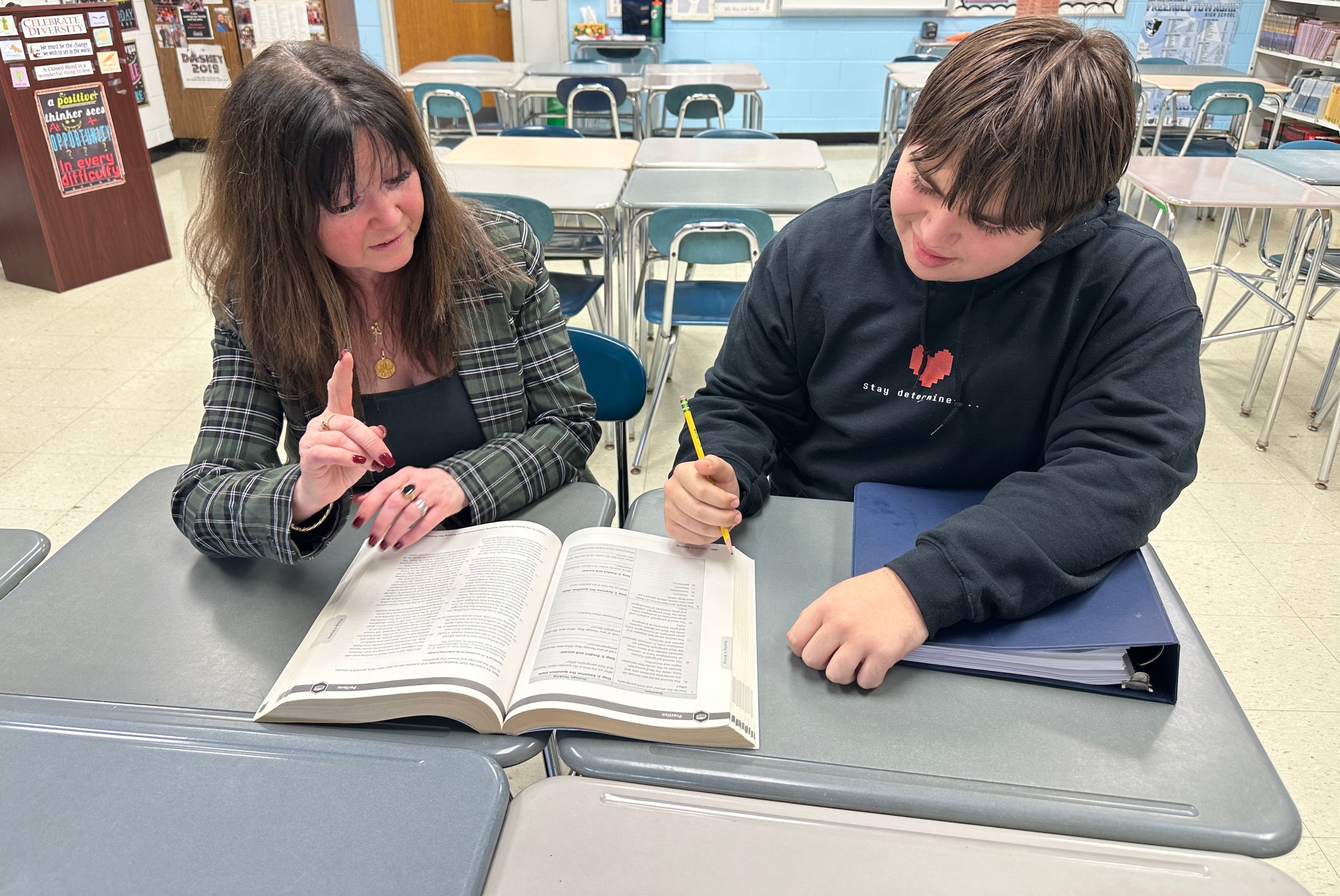 A woman and a teenage boy sitting at a school desk, looking at an open book with the woman pointing at it. The classroom has blue and white walls, bookshelves, and empty desks in the background.