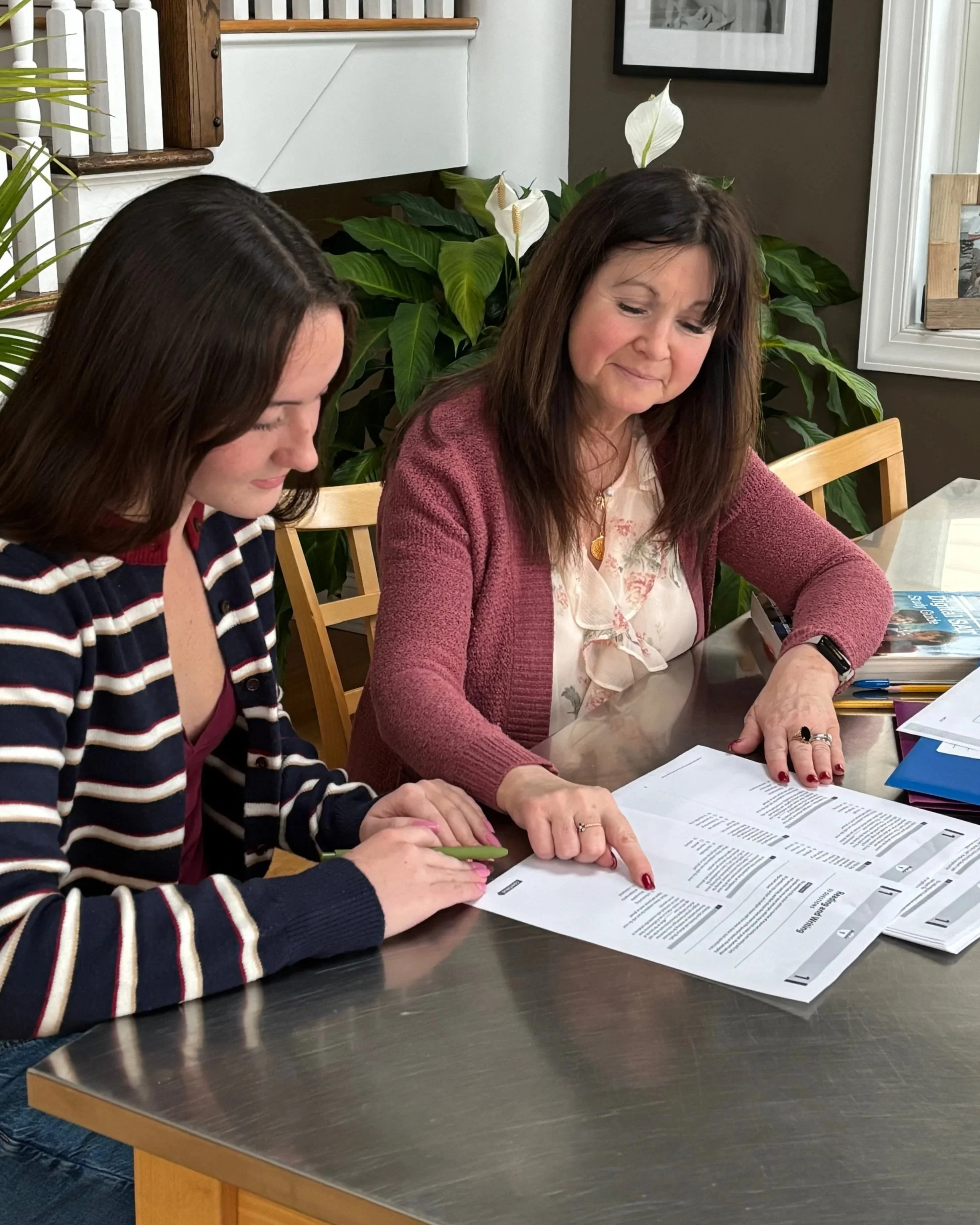 Two women sitting at a table looking over documents. One woman is pointing at the papers while the other observes. There are plants and picture frames in the background.