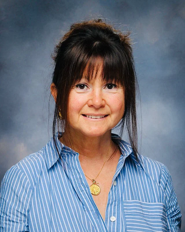 A woman with dark hair styled in an updo, wearing a blue and white striped button-down shirt and a gold necklace, smiling at the camera against a blue-gray background.