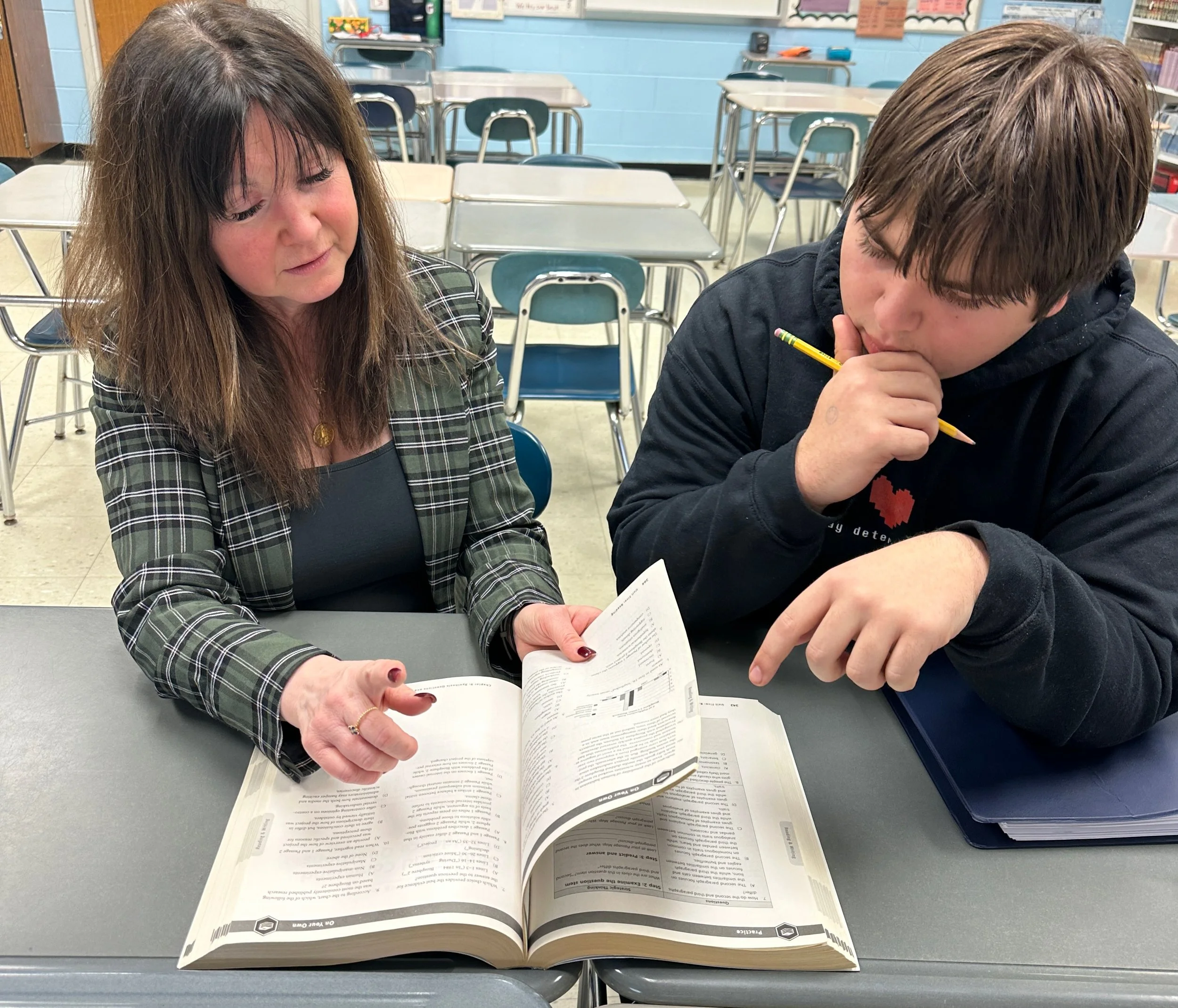 A woman and a teenage boy sitting at a school desk in a classroom, looking at an open textbook together. The woman is pointing to a section in the book, and the boy is holding a pencil near his mouth, appearing to be thinking.