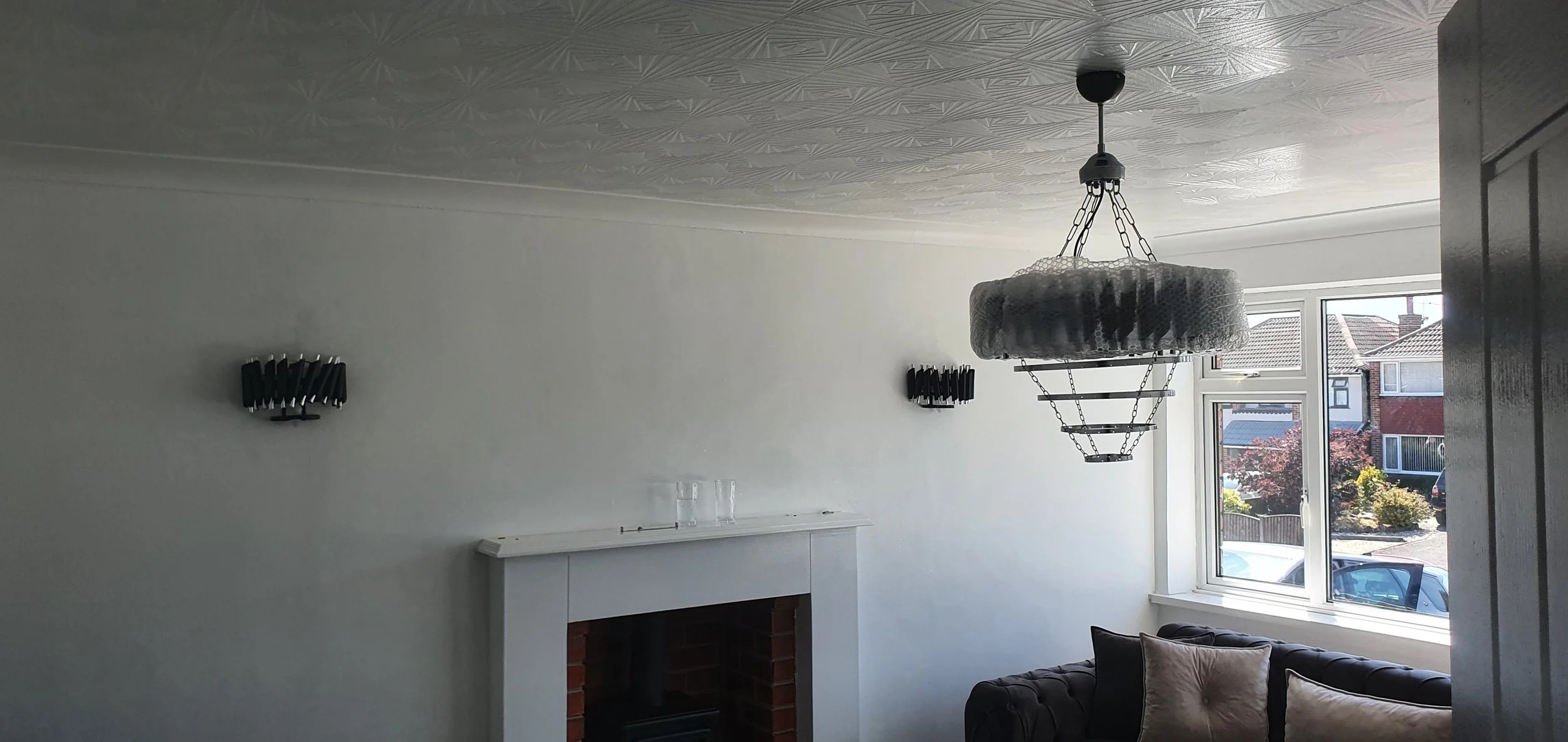 Living room with a white wall, decorative black sconces, a brick fireplace, and large windows showing a neighborhood outside. A chandelier hangs from the ceiling, and a black sofa with beige pillows is in the corner.