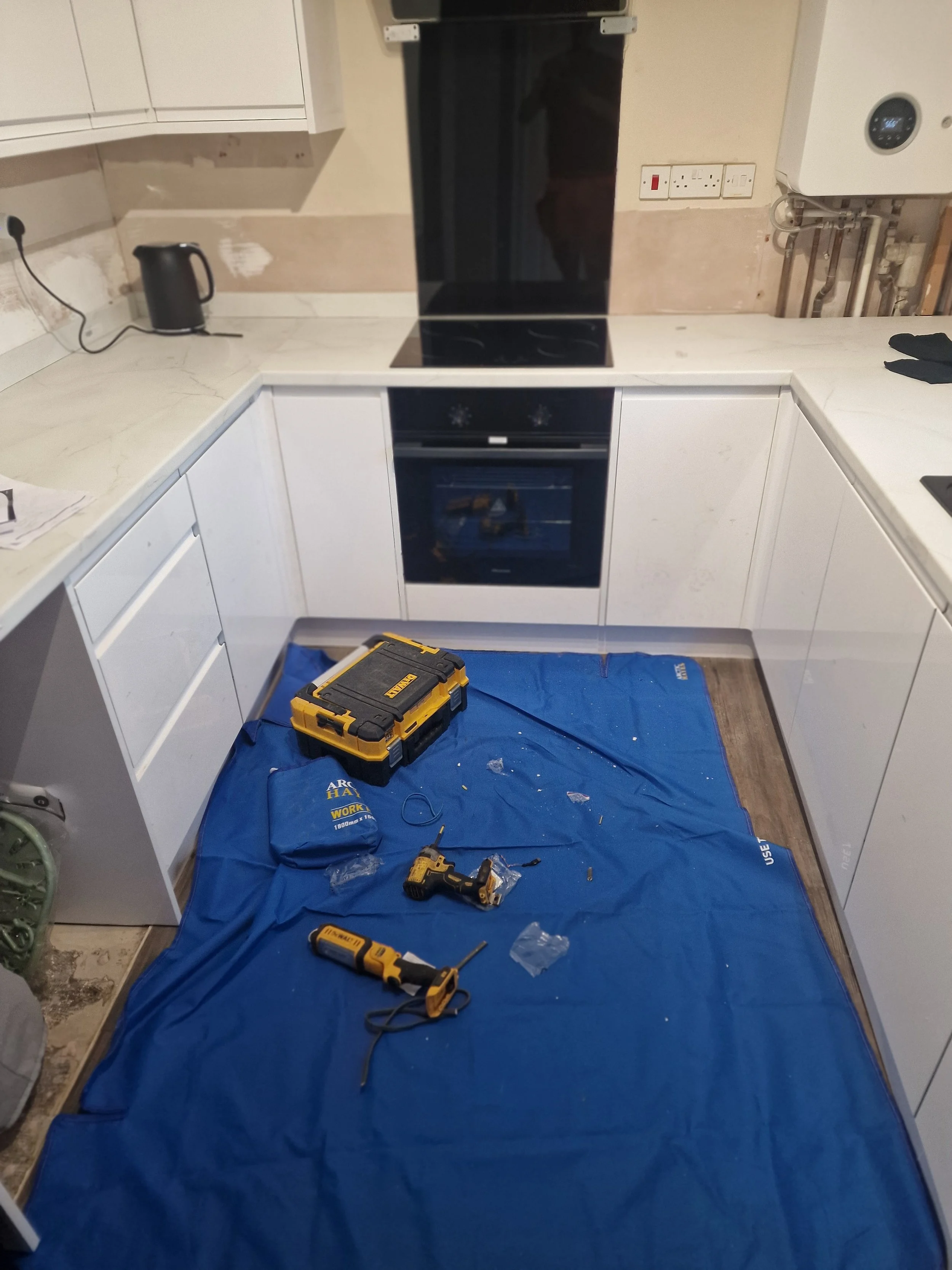 Kitchen with countertops and cabinets, tools and equipment on the floor covered with a blue tarp, including a yellow toolbox, cordless drill, and other tools, suggesting ongoing renovation or repair work.