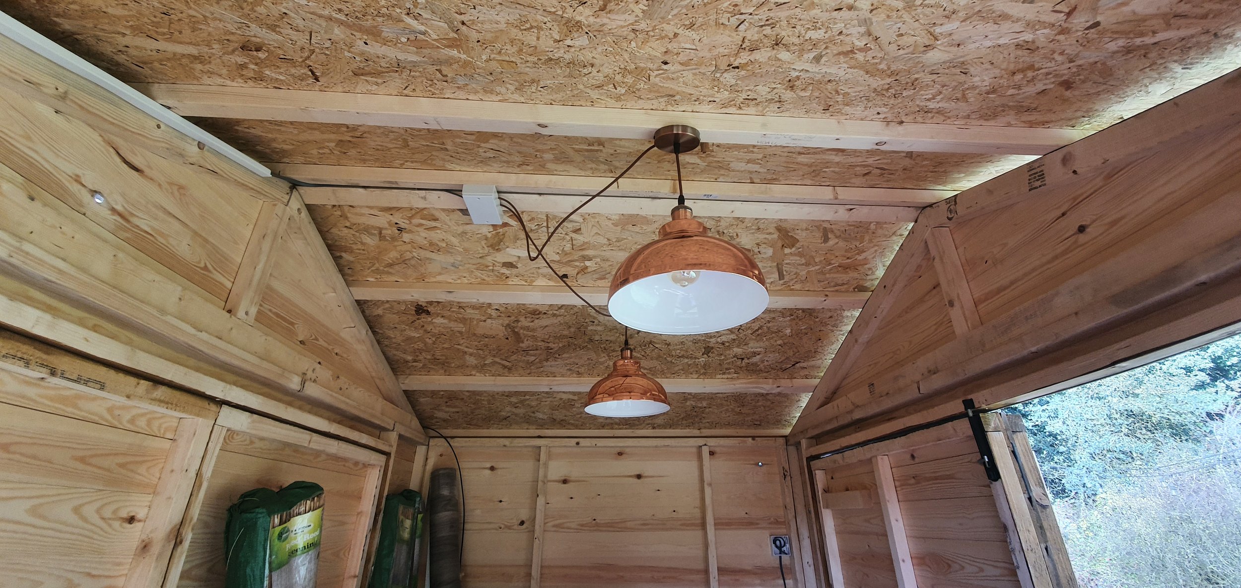 Interior of a wooden shed under construction with exposed plywood walls and ceiling, two hanging copper-colored pendant lights, and a window showing trees outside.