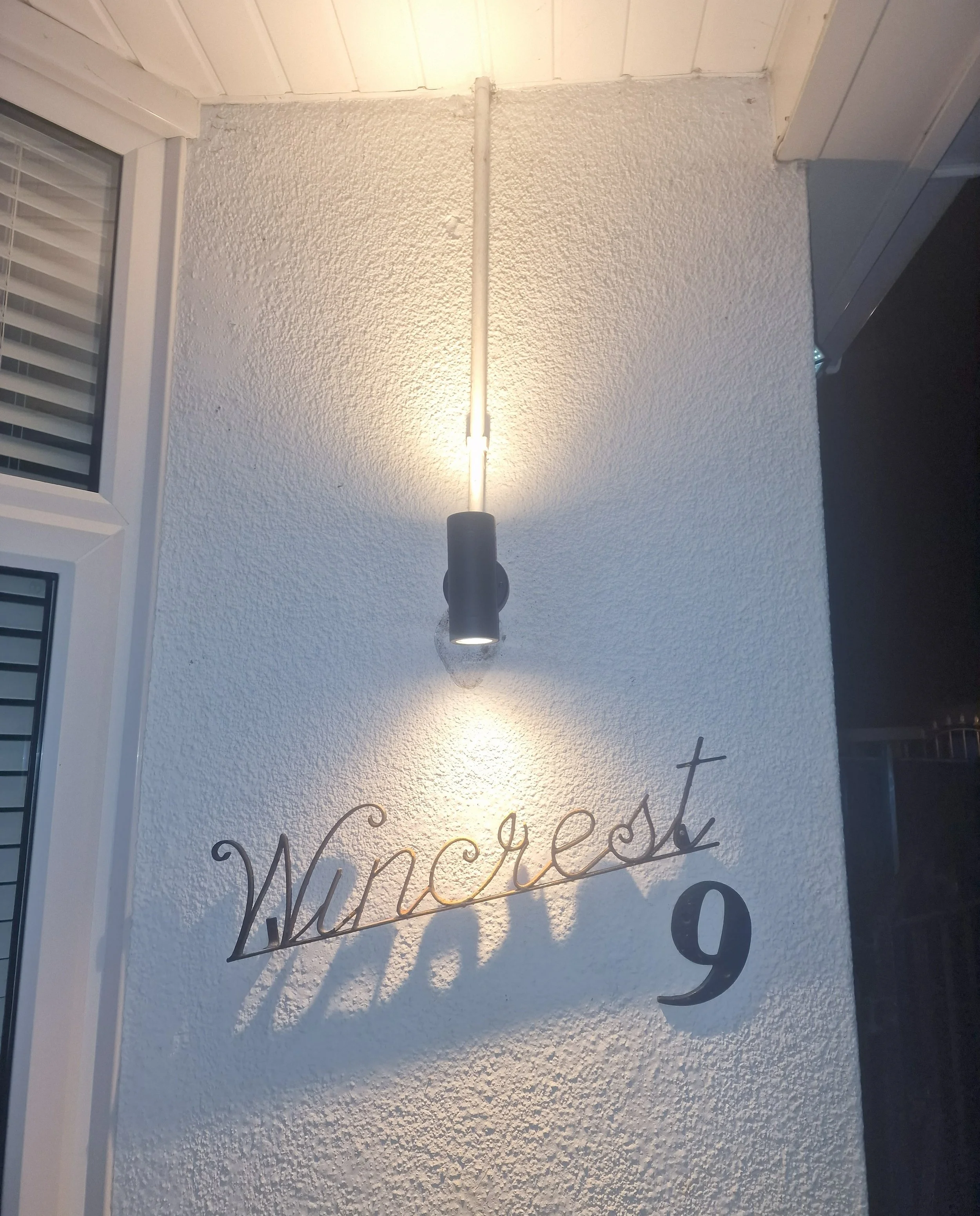Exterior wall with a spotlight, house number 9, and the word 'Wincrest' in decorative script. A window with shutters is visible on the left.