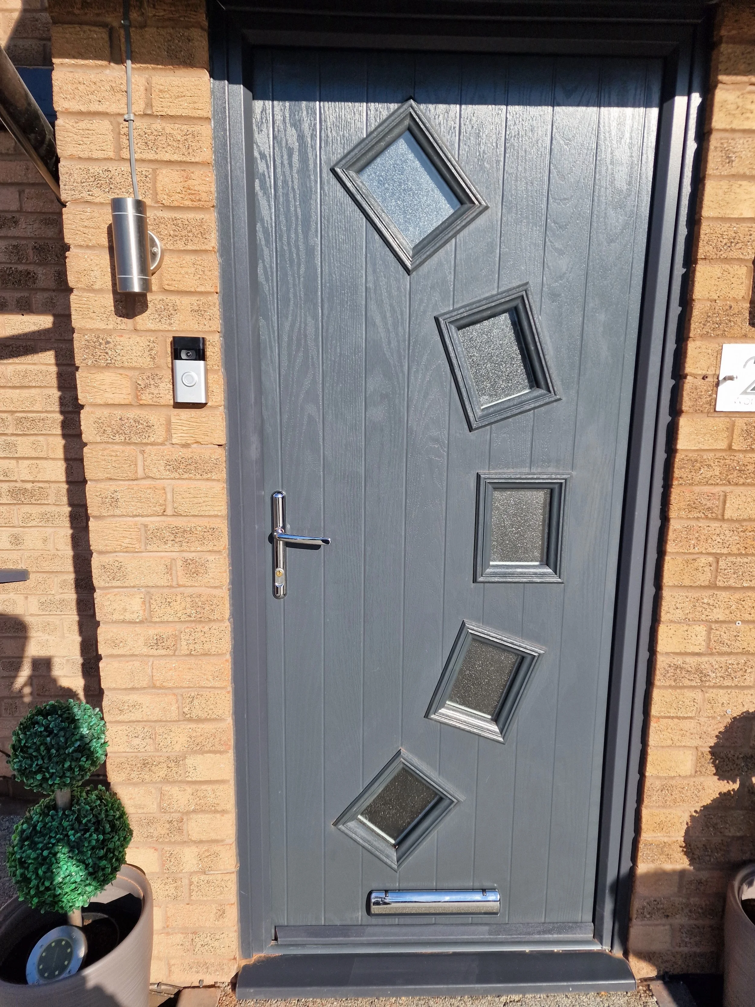 Gray front door with four tilted diamond-shaped windows, brick wall, door handle, mail slot, and outdoor light fixture.