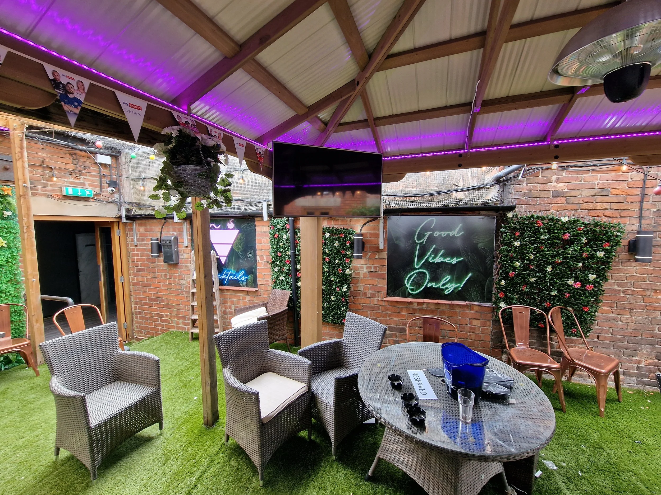 Indoor patio with artificial grass flooring, wicker chairs around a glass table, potted plants, flamingo sign, neon sign saying 'Good Vibes Only!', and pink and white flowers on brick walls, decorated with string lights.