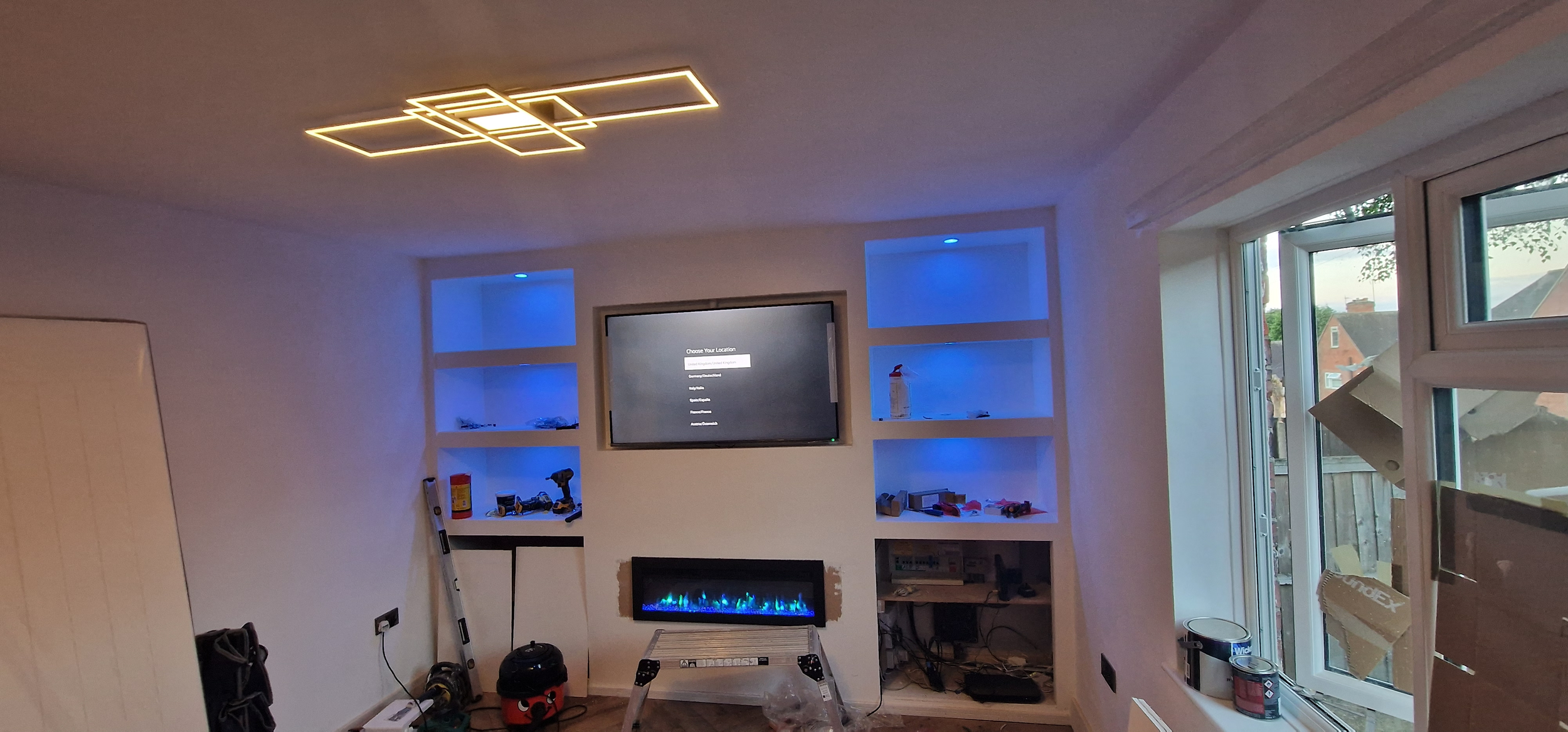 Living room with a modern ceiling light fixture, a large window, and a wall-mounted flat-screen TV above a faux fireplace with colorful LED lights. Built-in shelves on either side of the TV contain tools, a fire extinguisher, and various objects. The