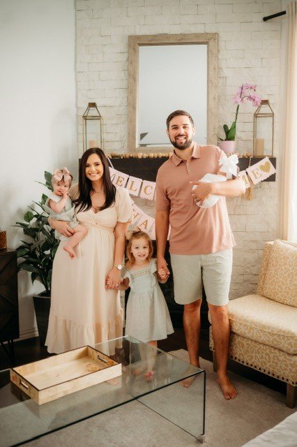 Family of four, including two young girls and two adults, standing in a living room decorated with pink flowers and banners, holding a newborn baby.