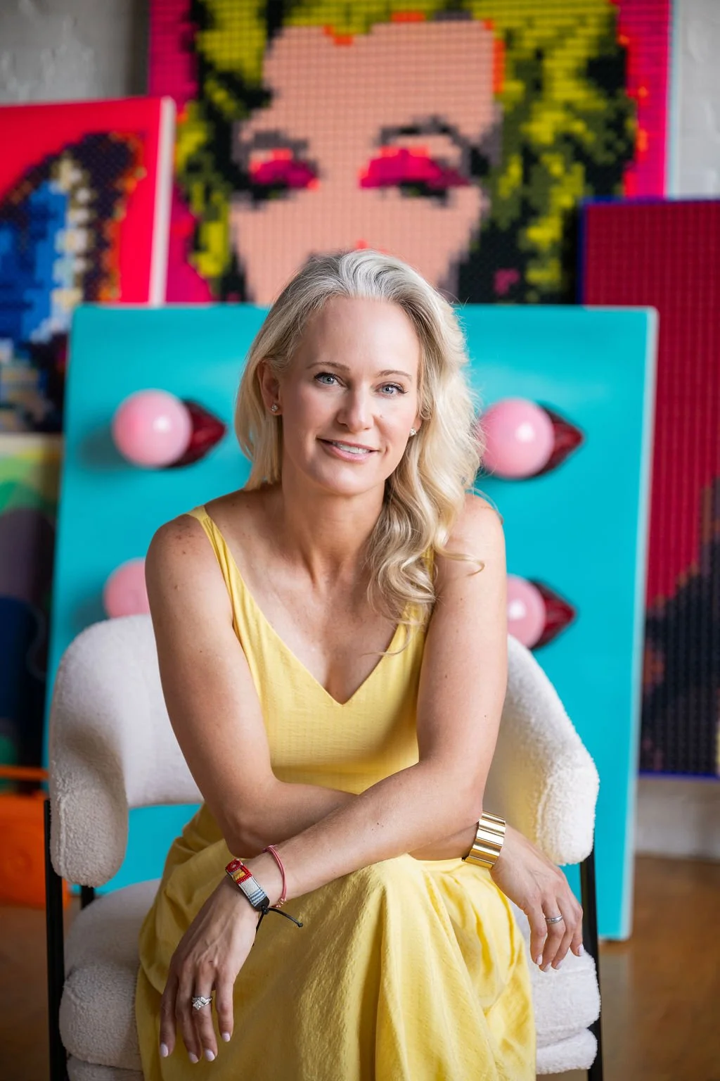A smiling woman with blonde wavy hair wearing a yellow sleeveless dress, sitting on a white armchair with a decorative background of colorful artwork including a large portrait of a woman's face with pink lips and eye makeup.