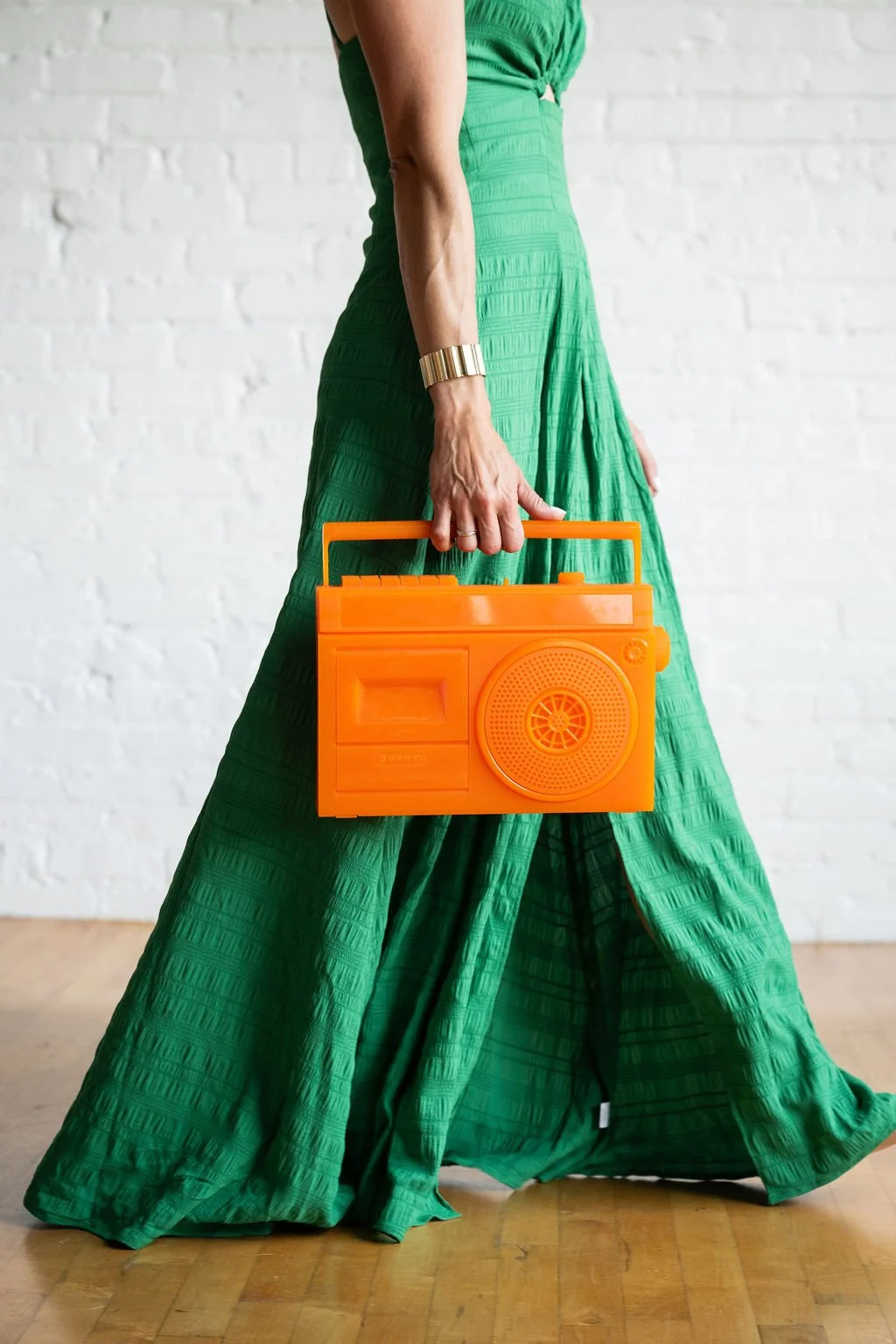 A person wearing a long green dress holding an orange boombox, standing on a wooden floor with a white brick wall in the background.