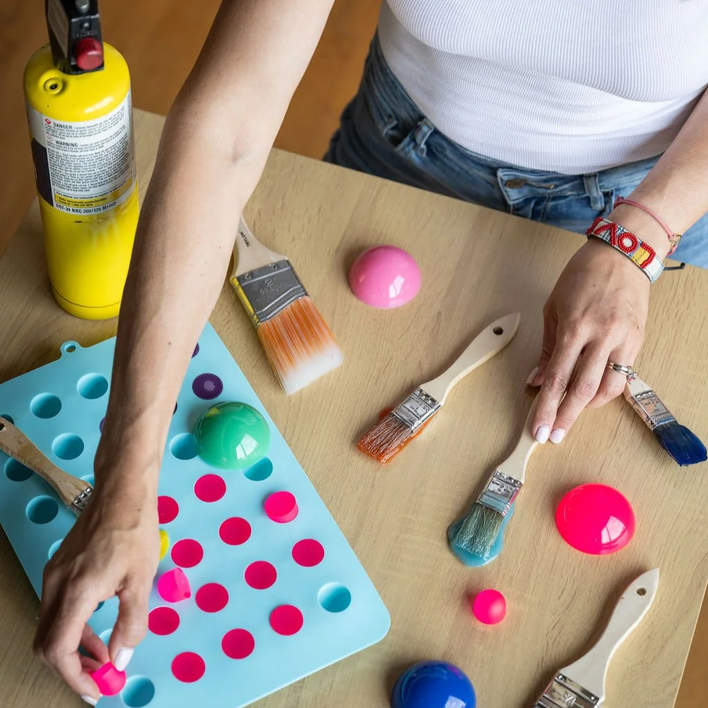 Person painting colorful objects with paint brushes on a wooden table. Paint containers and a silicone mold filled with pink paint are also visible.