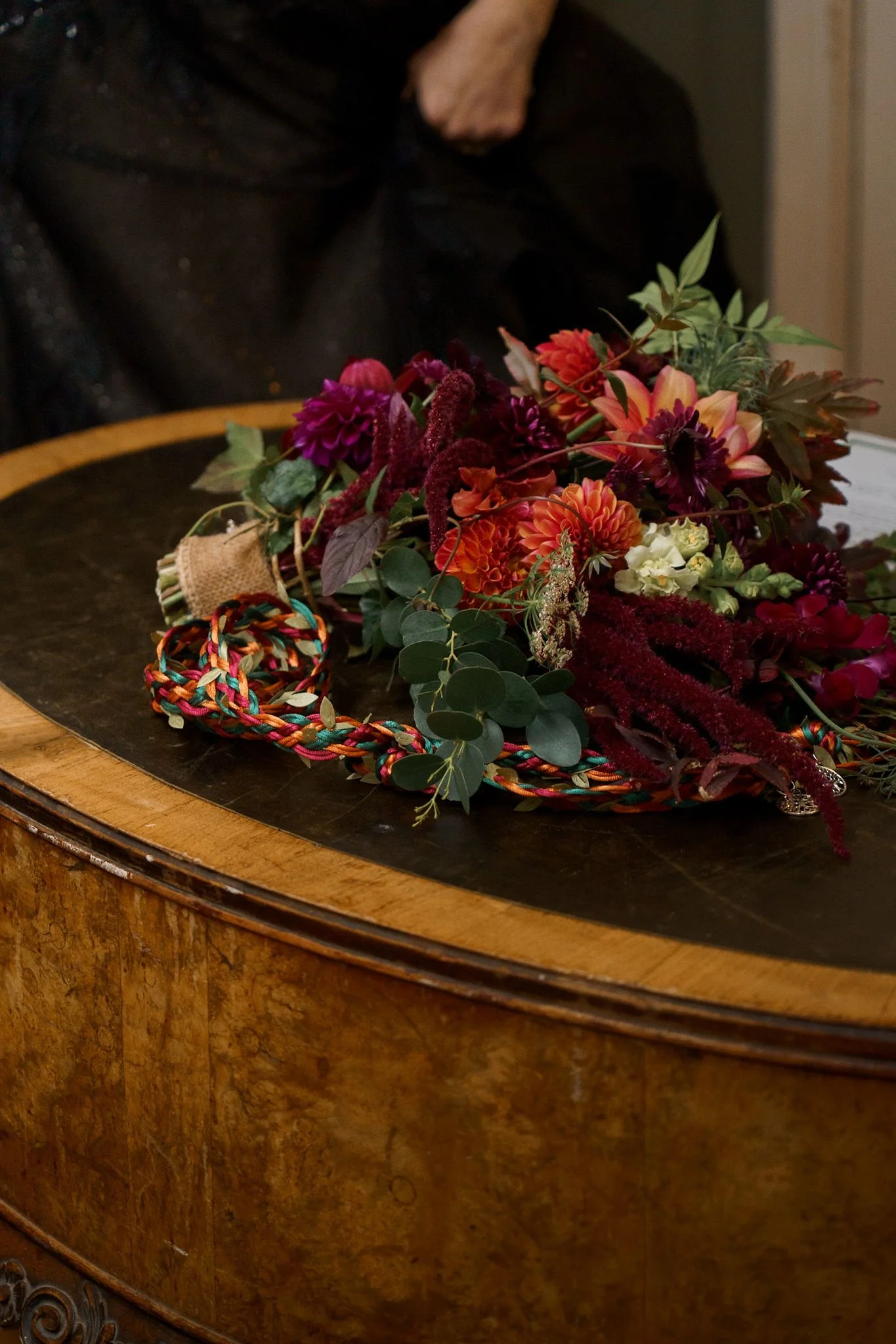 An autumnal wedding bouquet laid across a bespoke handfasting cord, atop a leather covered table used for signing documents