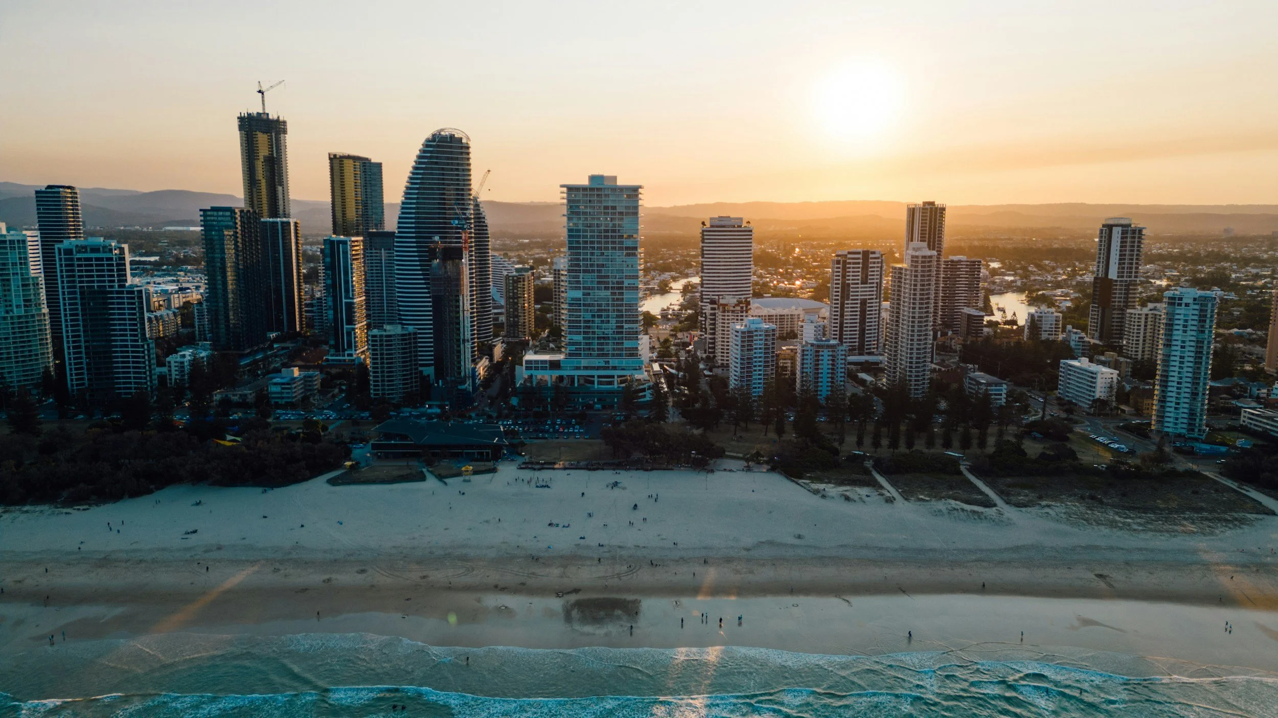 A city skyline at sunset with tall skyscrapers, a sandy beach in the foreground, and a river running through the city.