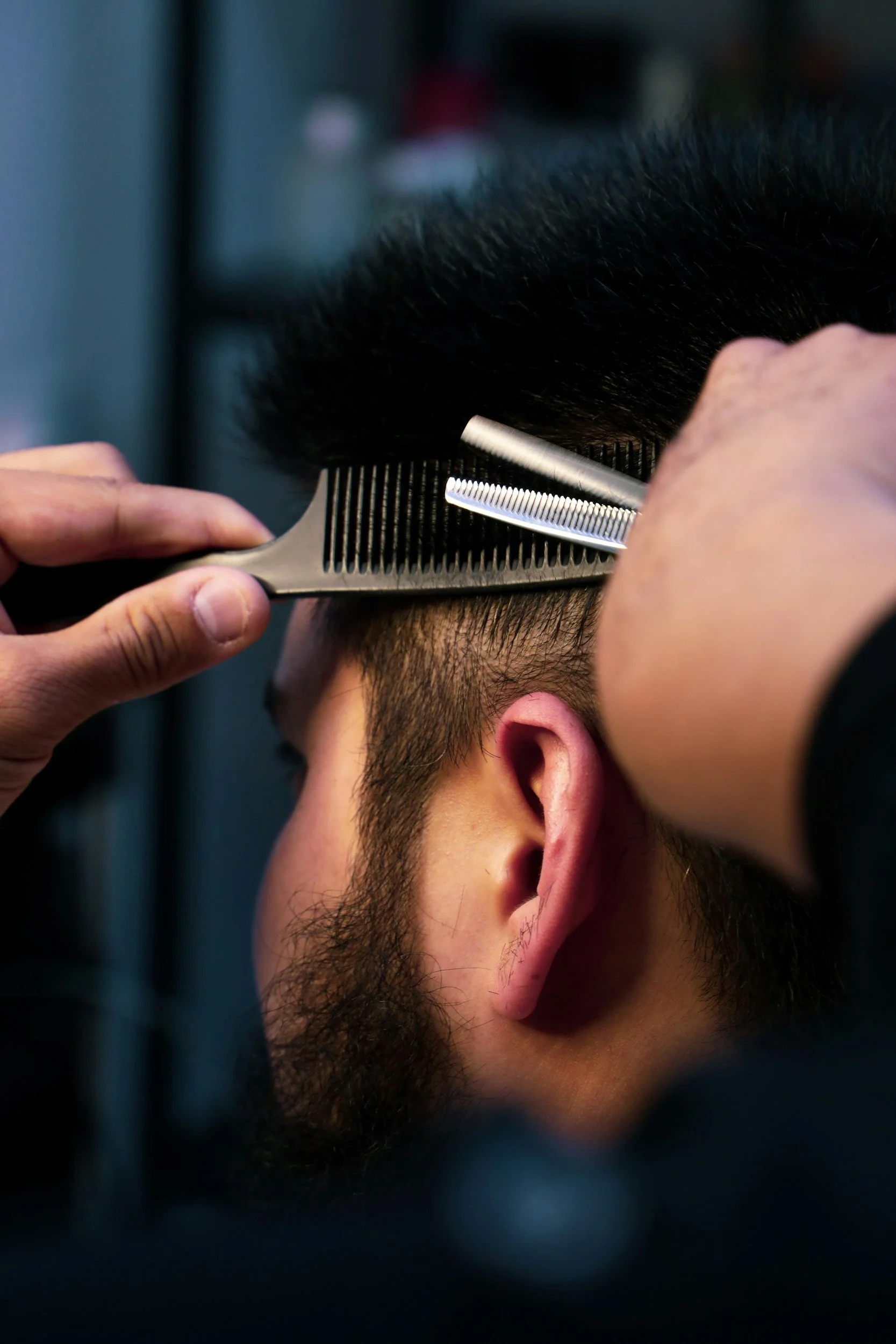 A person getting a haircut, with a barber trimming the hair with scissors and a comb, close-up view of the side of the person's head and ear.