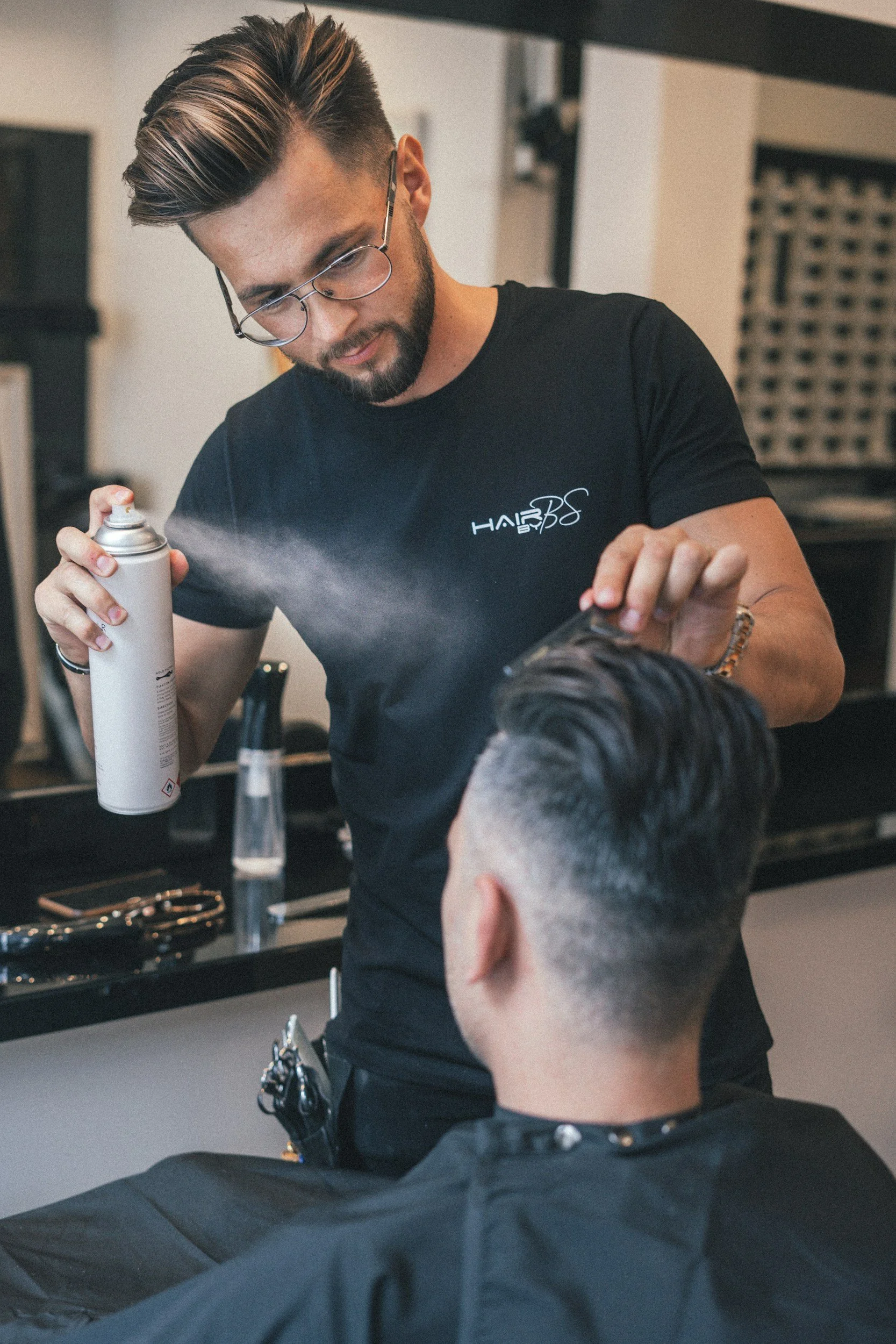 A hairstylist sprays hair product onto a male client's hair at a barbershop.
