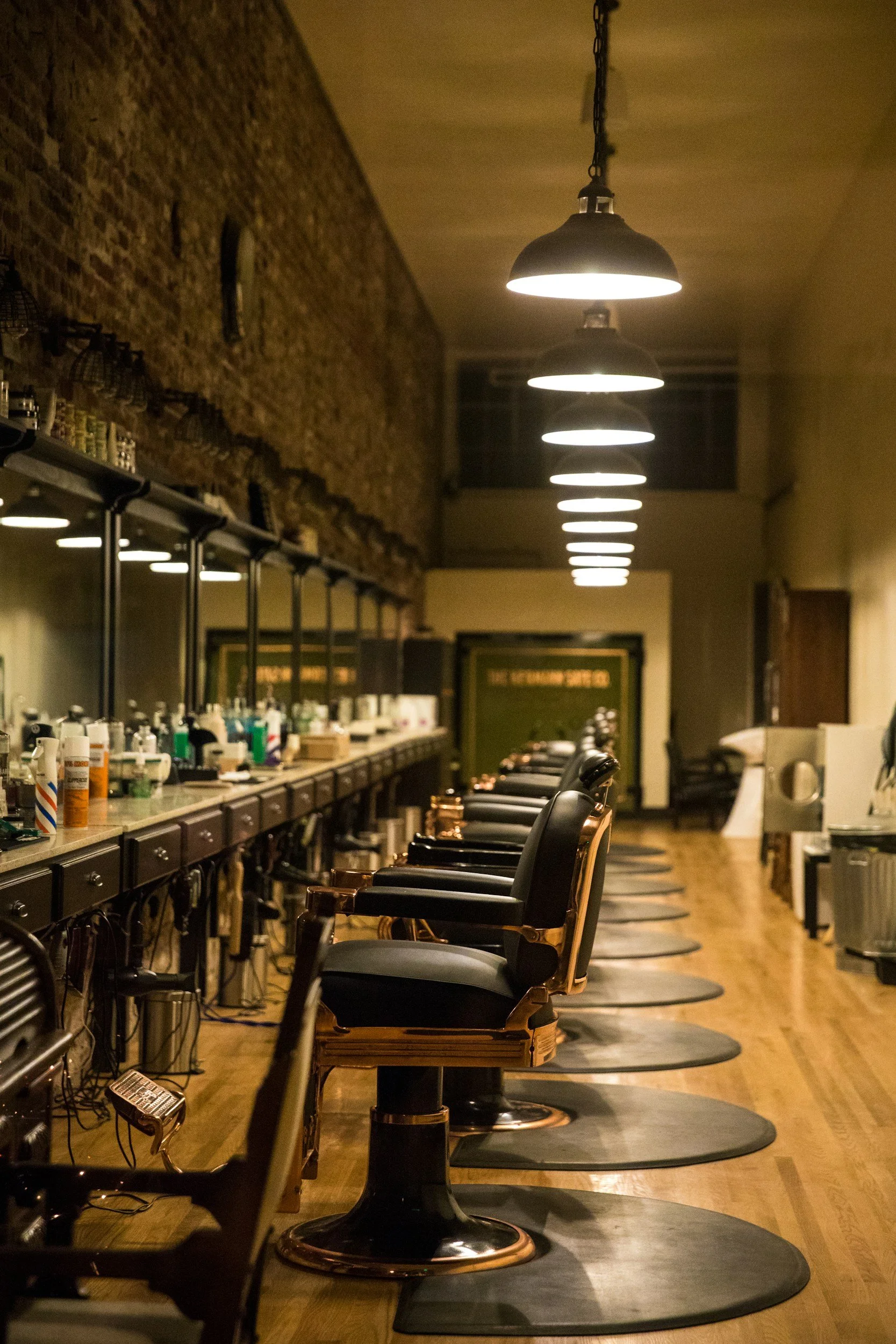 Empty barbershop with black and gold chairs lined up in front of mirrors, overhead lights, and barber supplies on the counter.
