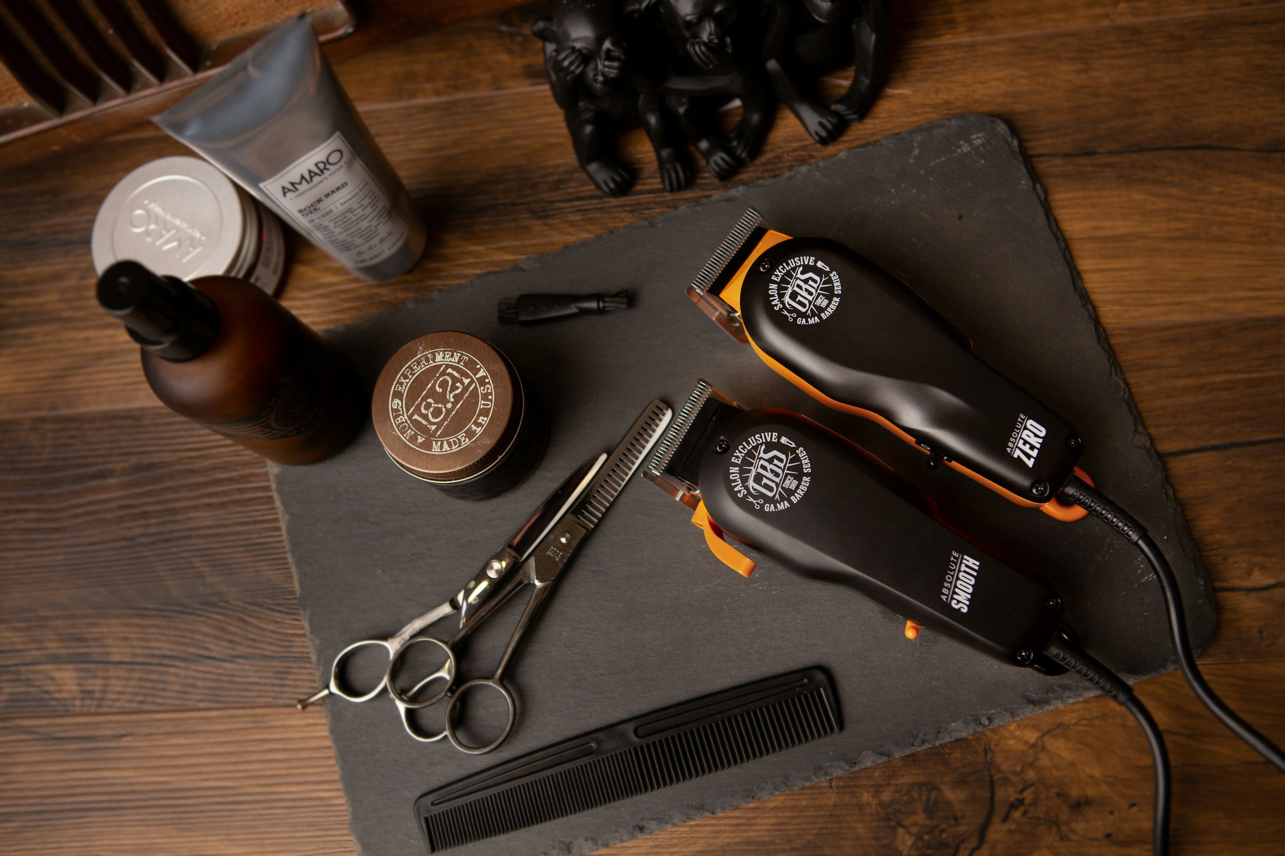 A flat lay of barber tools and grooming products on a dark slate surface, including hair clippers, scissors, a hair comb, bottles, and containers.