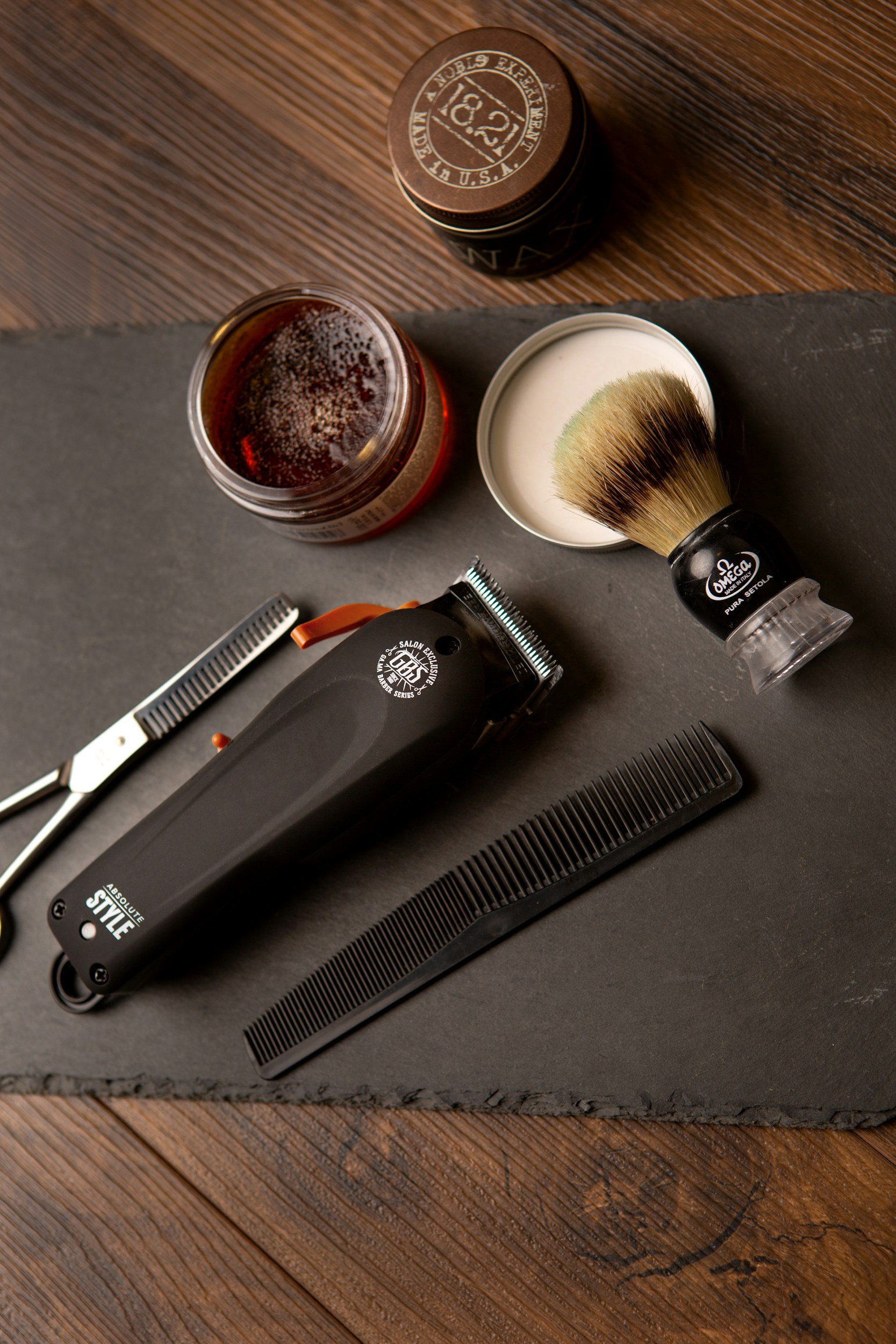 Barber tools including a hair clipper, a comb, a jar of shaving cream, a shaving brush, and grooming products on a dark slate surface.