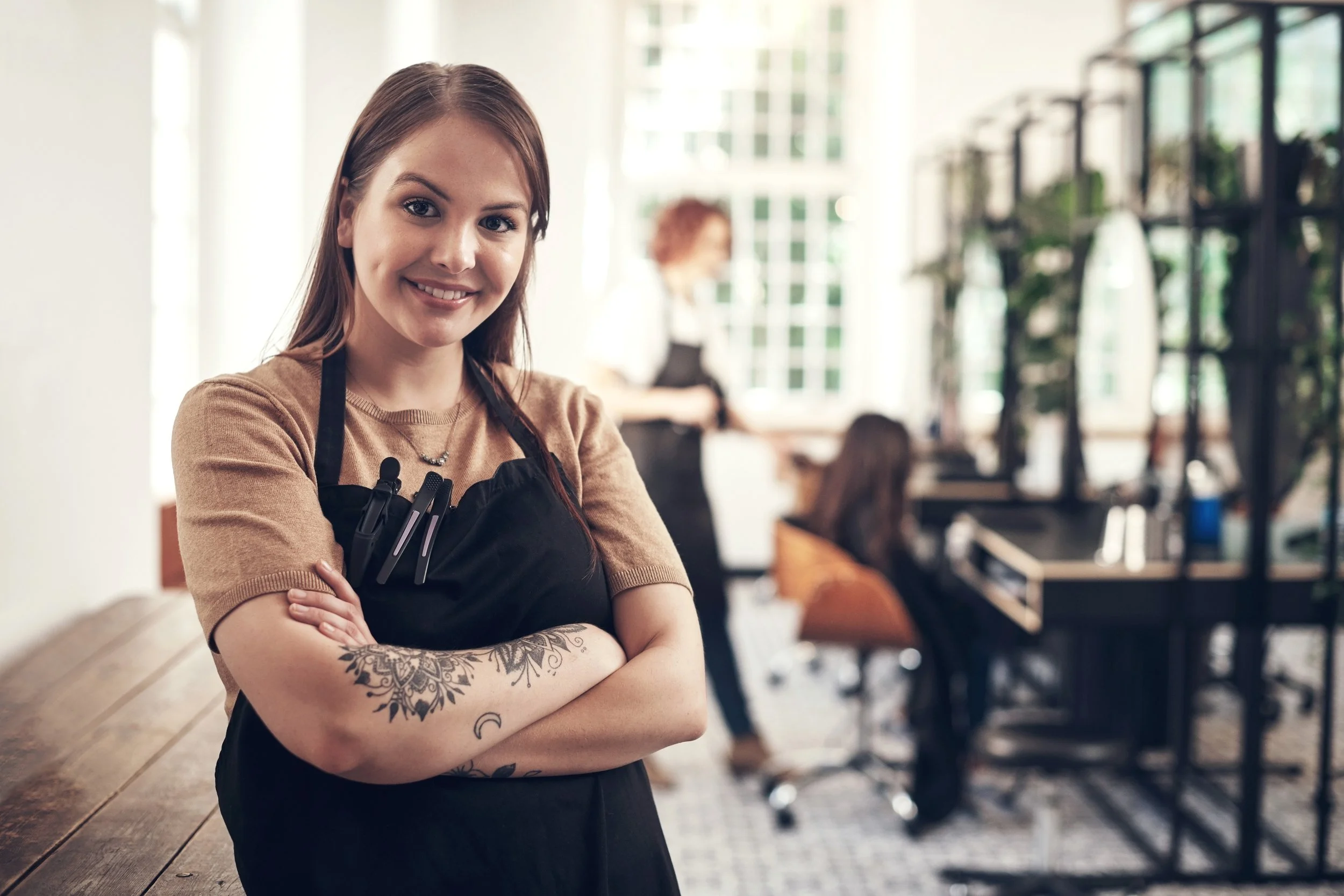 Smiling young woman with arms crossed, wearing a black apron with pens, inside a well-lit modern salon with hairdressing equipment and a workstation in the background.