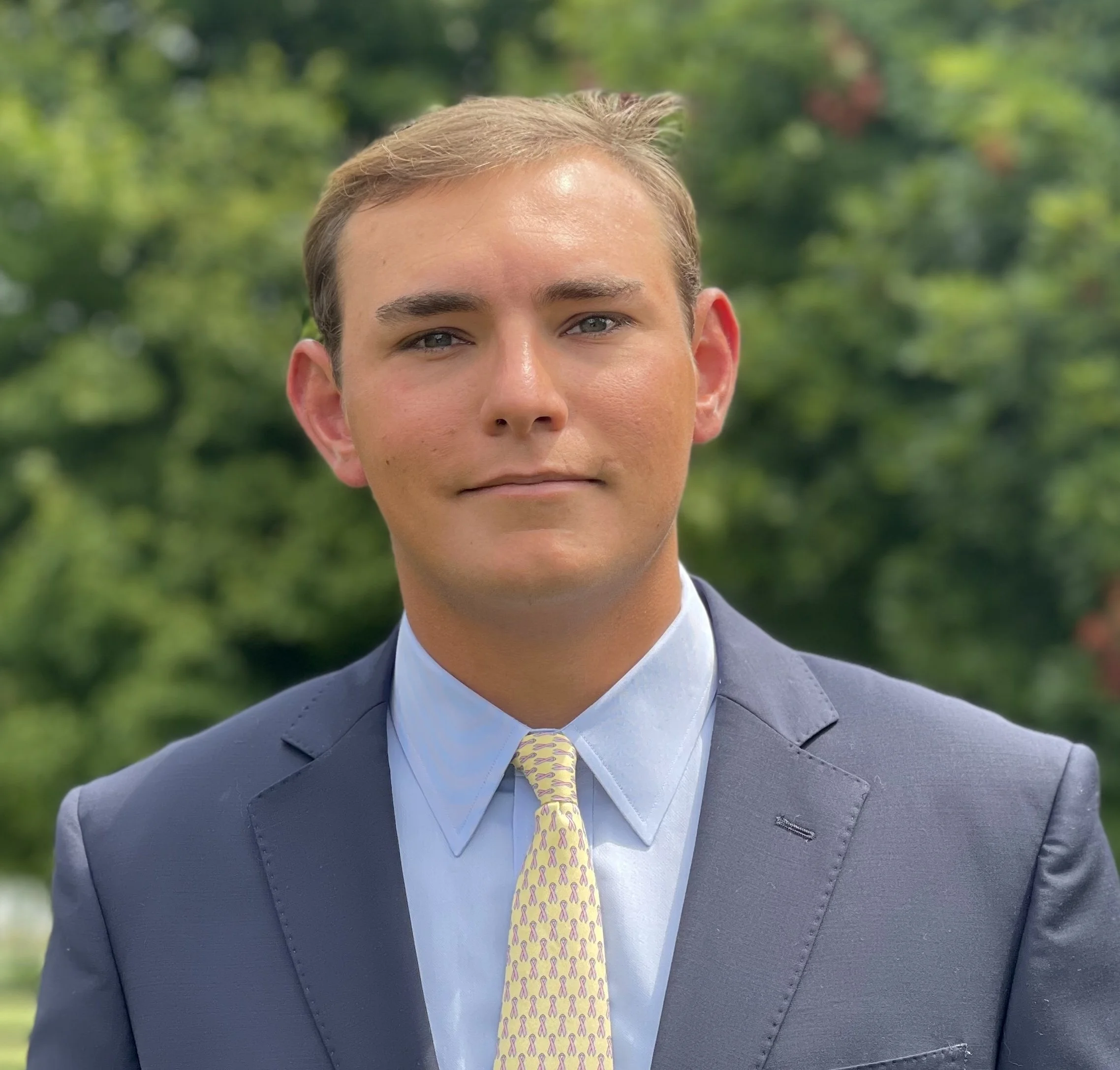 A man in a navy suit, light blue shirt, and a yellow patterned tie standing outdoors with greenery in the background.