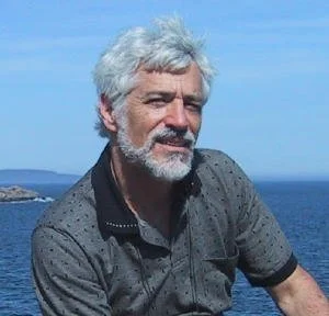 A man with gray hair and a beard, wearing a gray short-sleeved shirt, standing outdoors near the ocean.