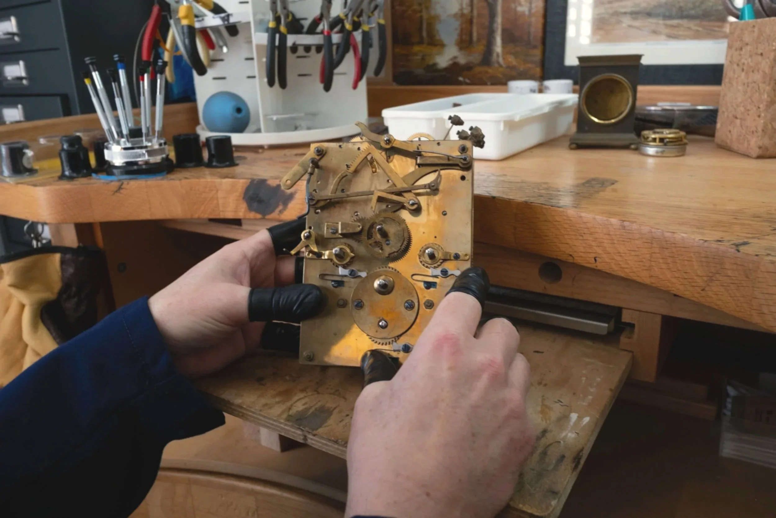 Person wearing black gloves holding an internal mechanical clockwork mechanism in a woodworking or repair workshop.