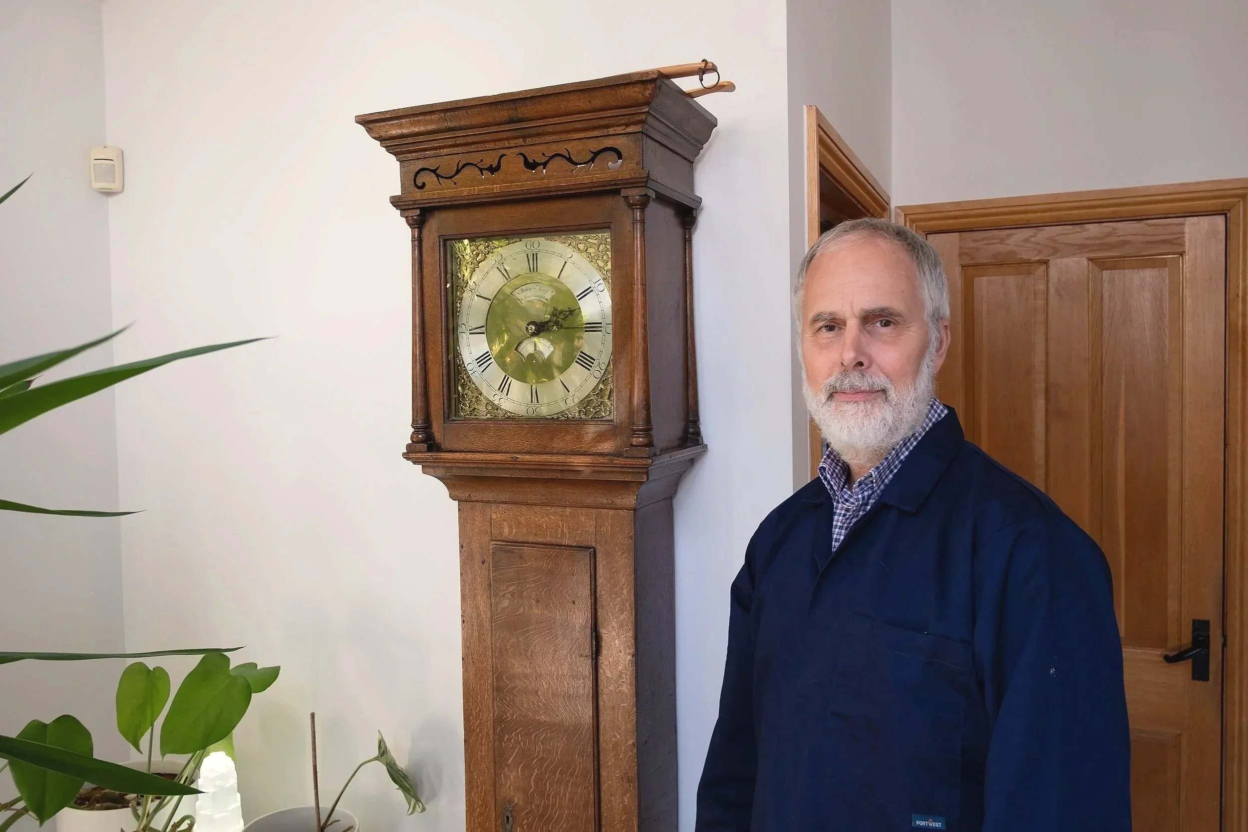 An older man with a gray beard wearing a dark jacket standing next to a tall antique wooden clock in a room with light-colored walls and a closed wooden door.