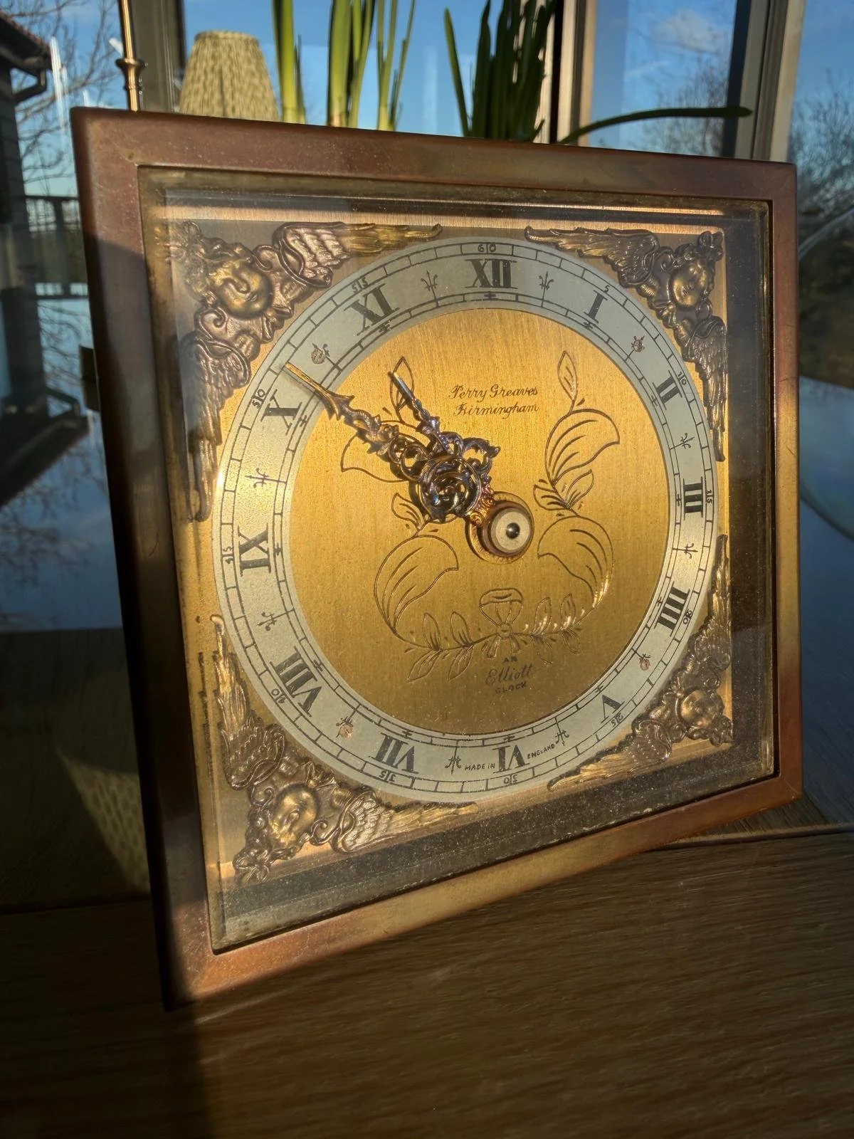 Vintage square wooden clock with ornate gold and silver detailing, Roman numerals, and a floral design on the face, placed on a wooden surface near a window with sunlight and a view of trees outside.