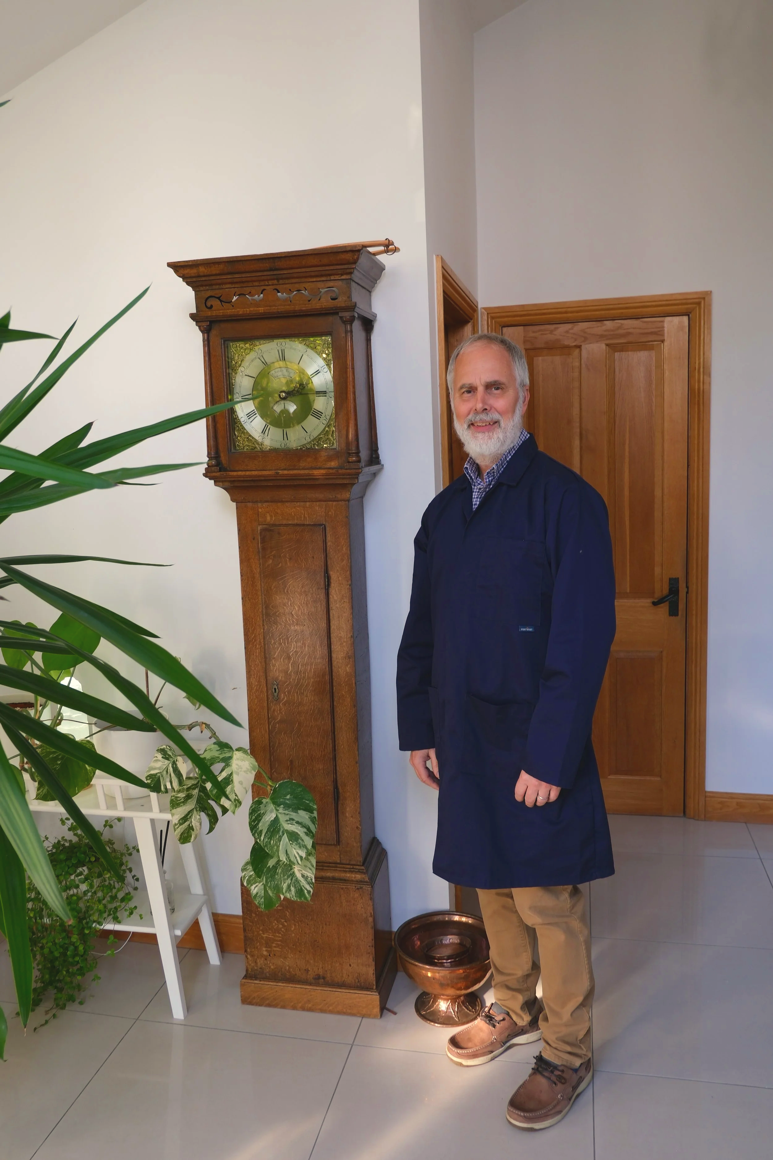 A man with gray hair and beard standing near a tall antique wooden clock and a copper bowl, in a room with white walls and wooden door frames, next to green houseplants.