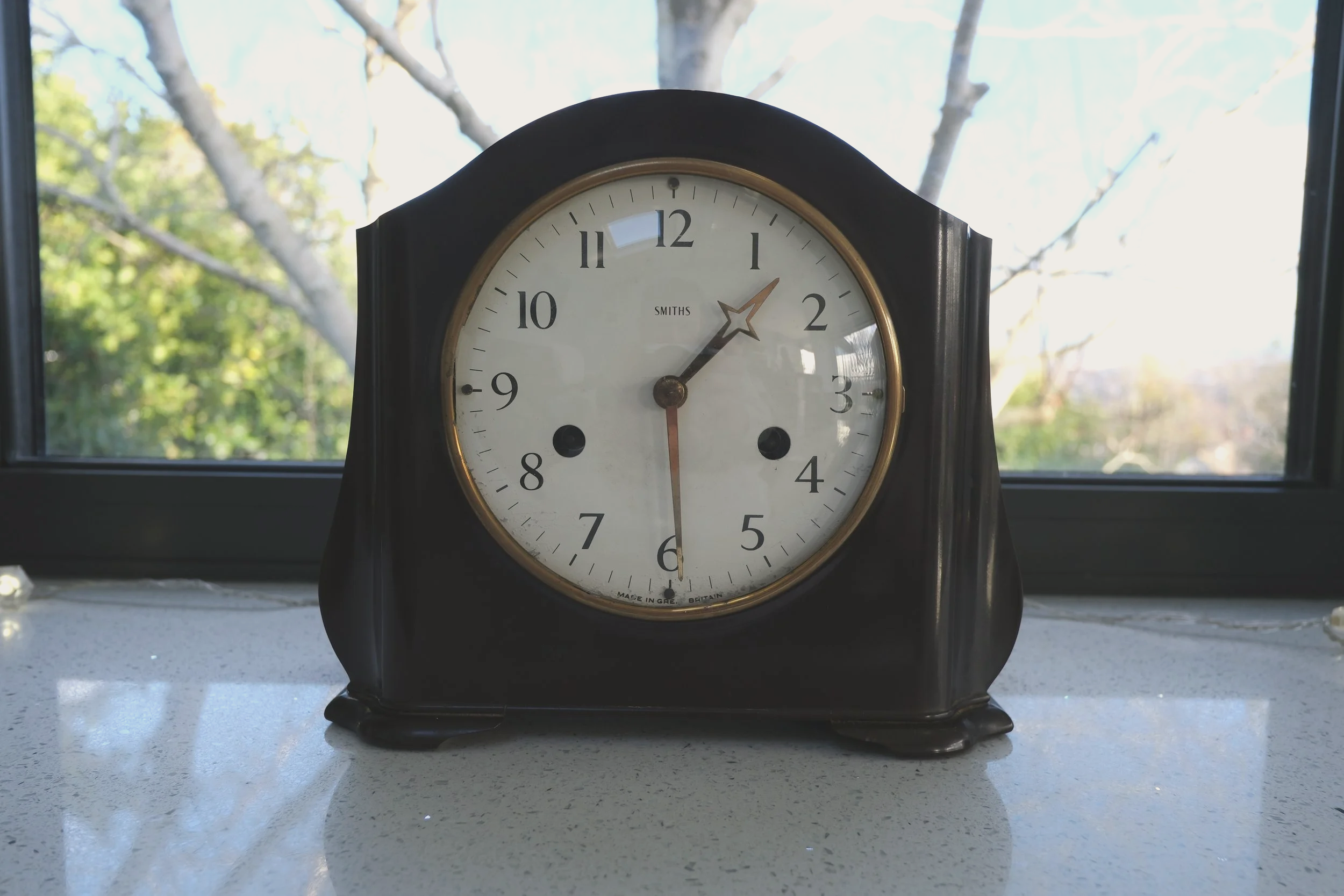 A vintage black and gold mantle clock showing the time as 1:29, placed on a white countertop in front of windows with a view of trees outside.