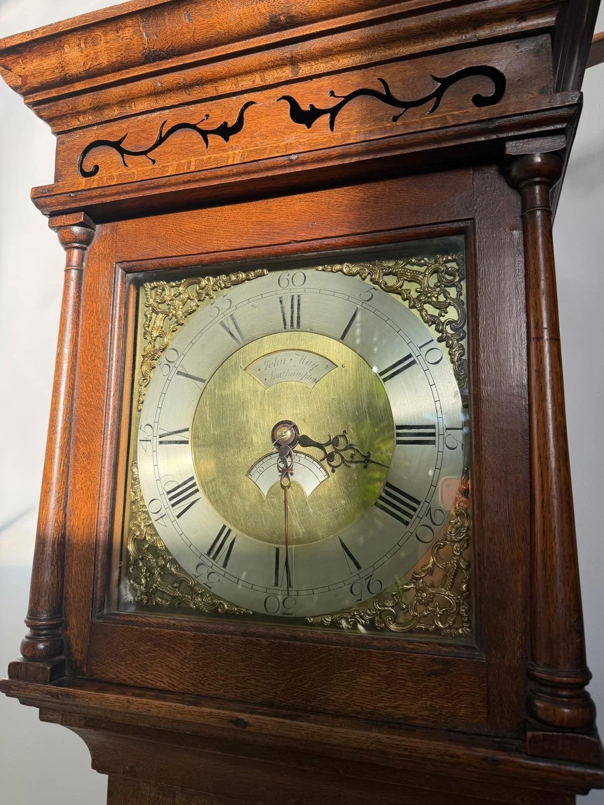 Close-up of an antique wooden clock with a brass dial, Roman numerals, intricate gold decorative details, and black hands, showing the time as 3:35.