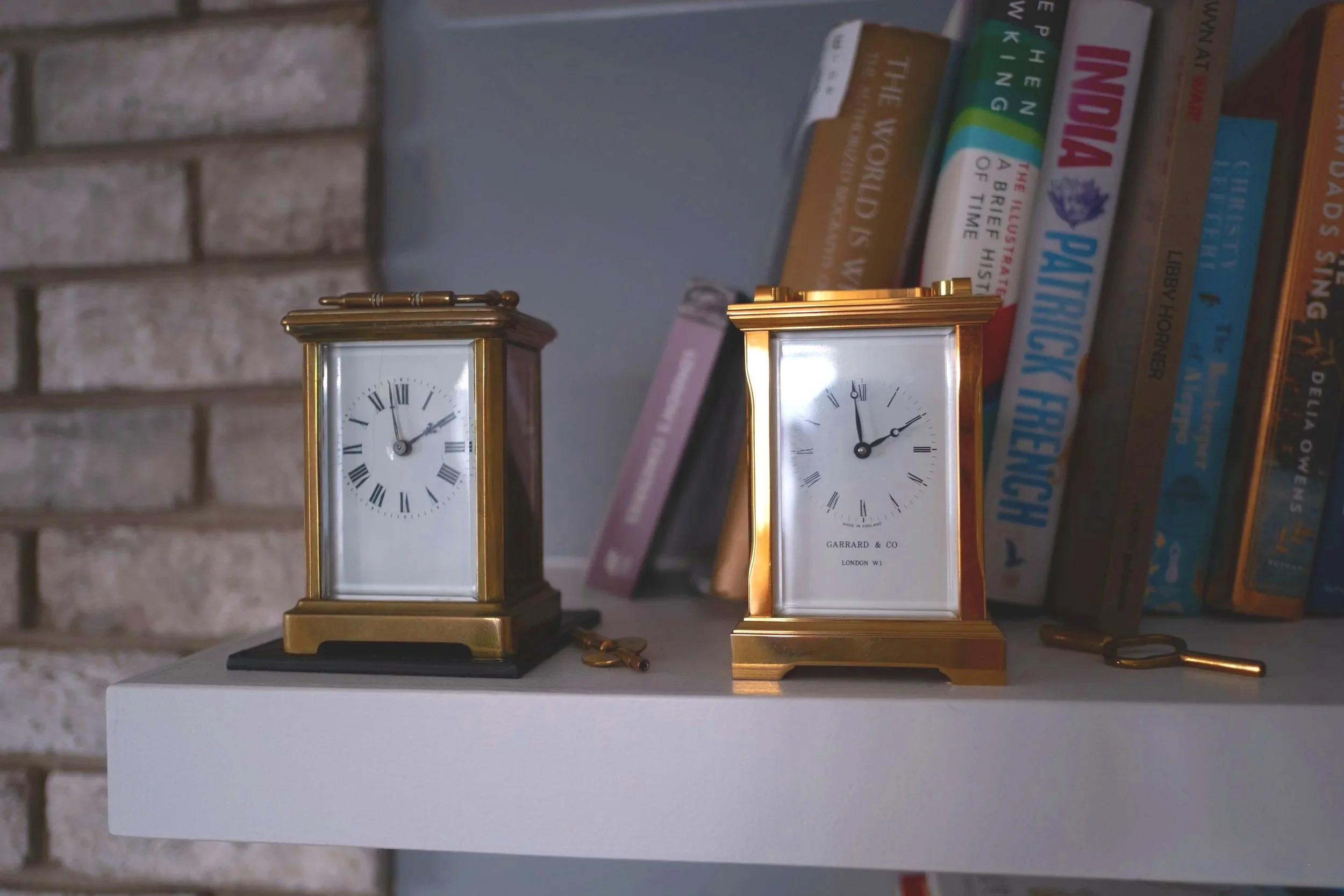 Two vintage tabletop clocks, one brass with a black base and the other gold, placed on a white shelf with books in the background. The clocks show different times, and there is a small key next to each clock.