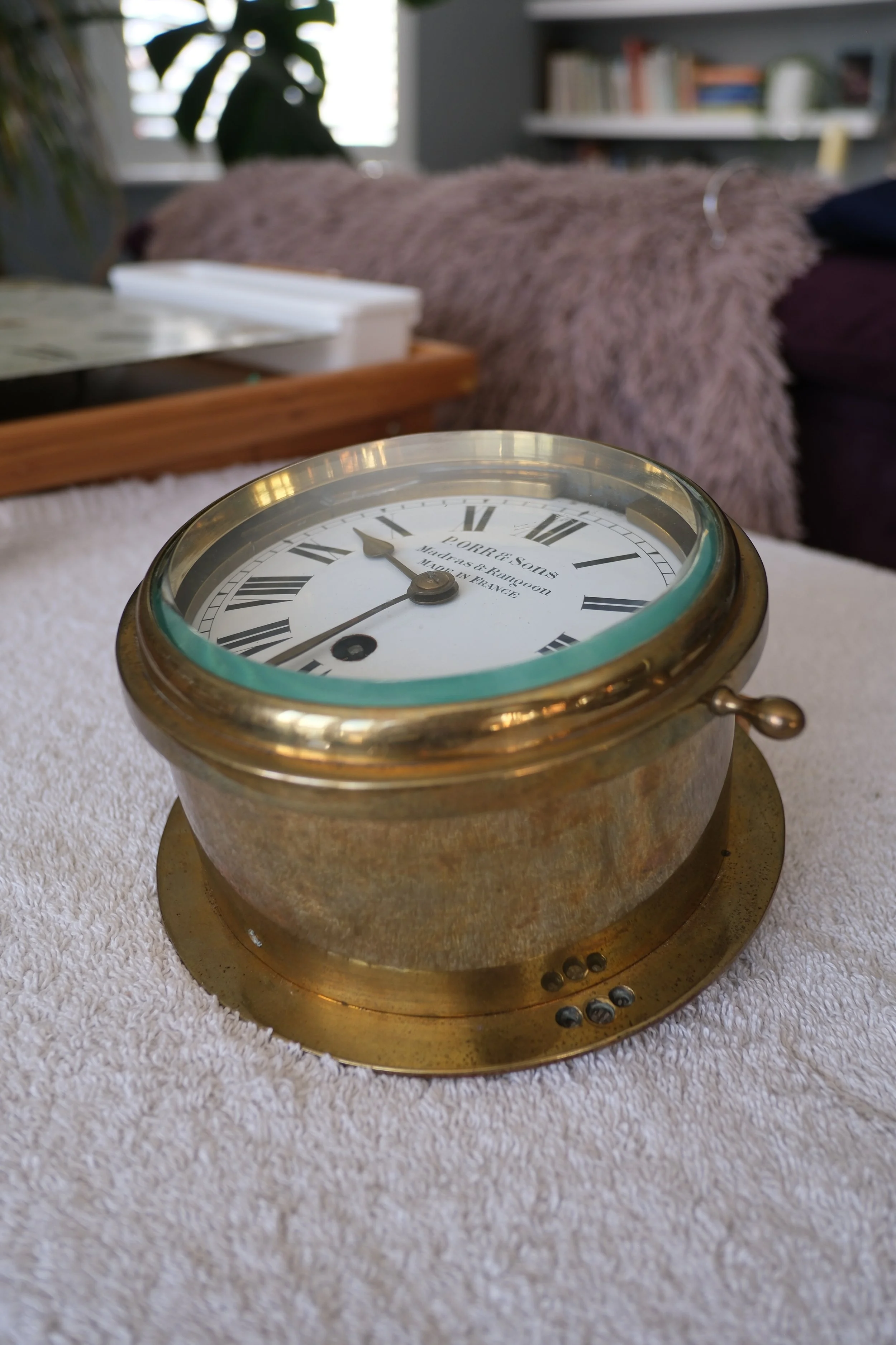 A vintage brass ship's clock on a white textured towel surface, with a blurred background including a bookshelf, a window, and furnishings.