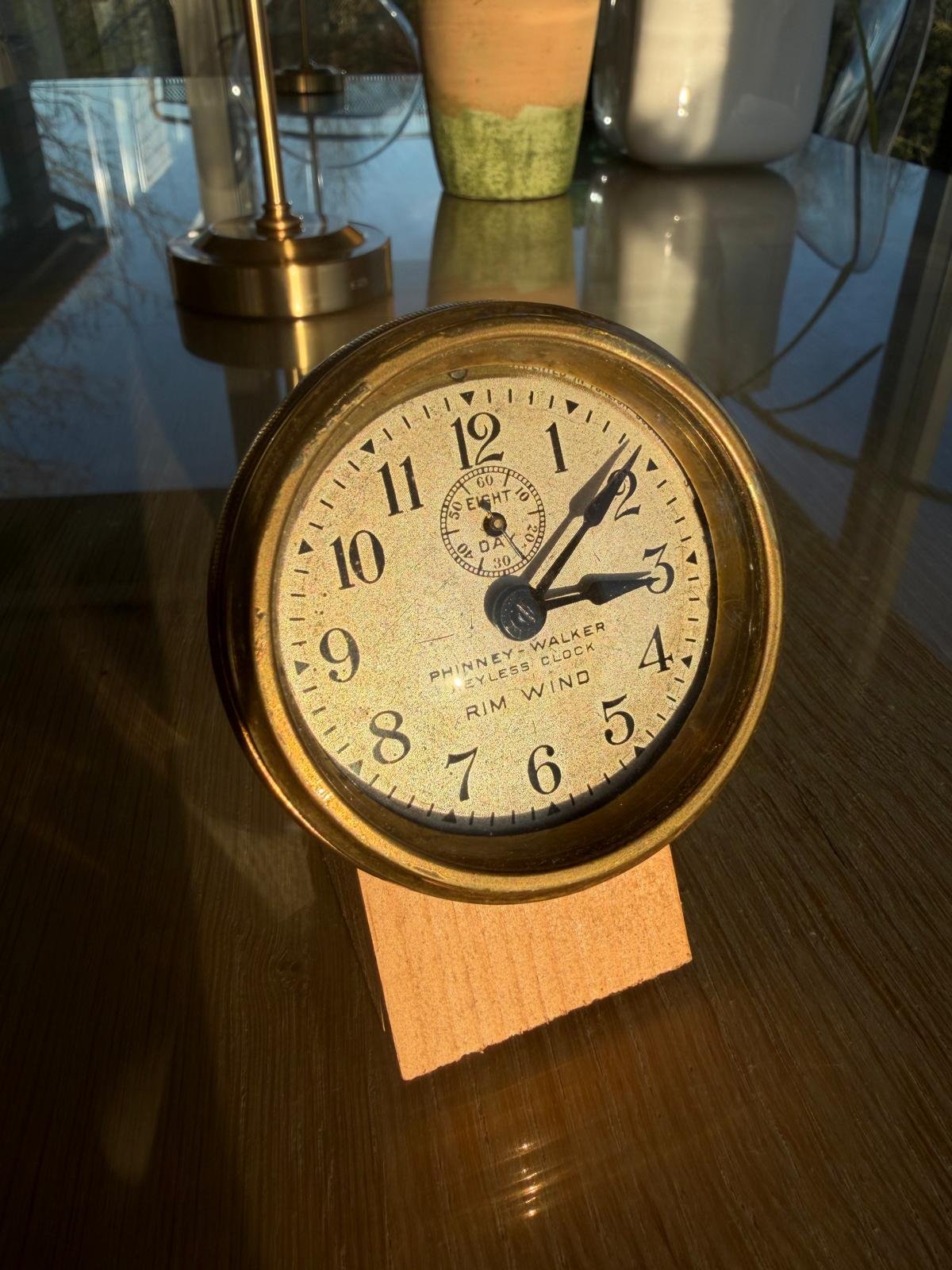 A vintage round clock displaying the time as 2:07, with a smaller dial at the top showing seconds, on a wooden stand on a reflective table with indoor lamps and potted plants in the background.