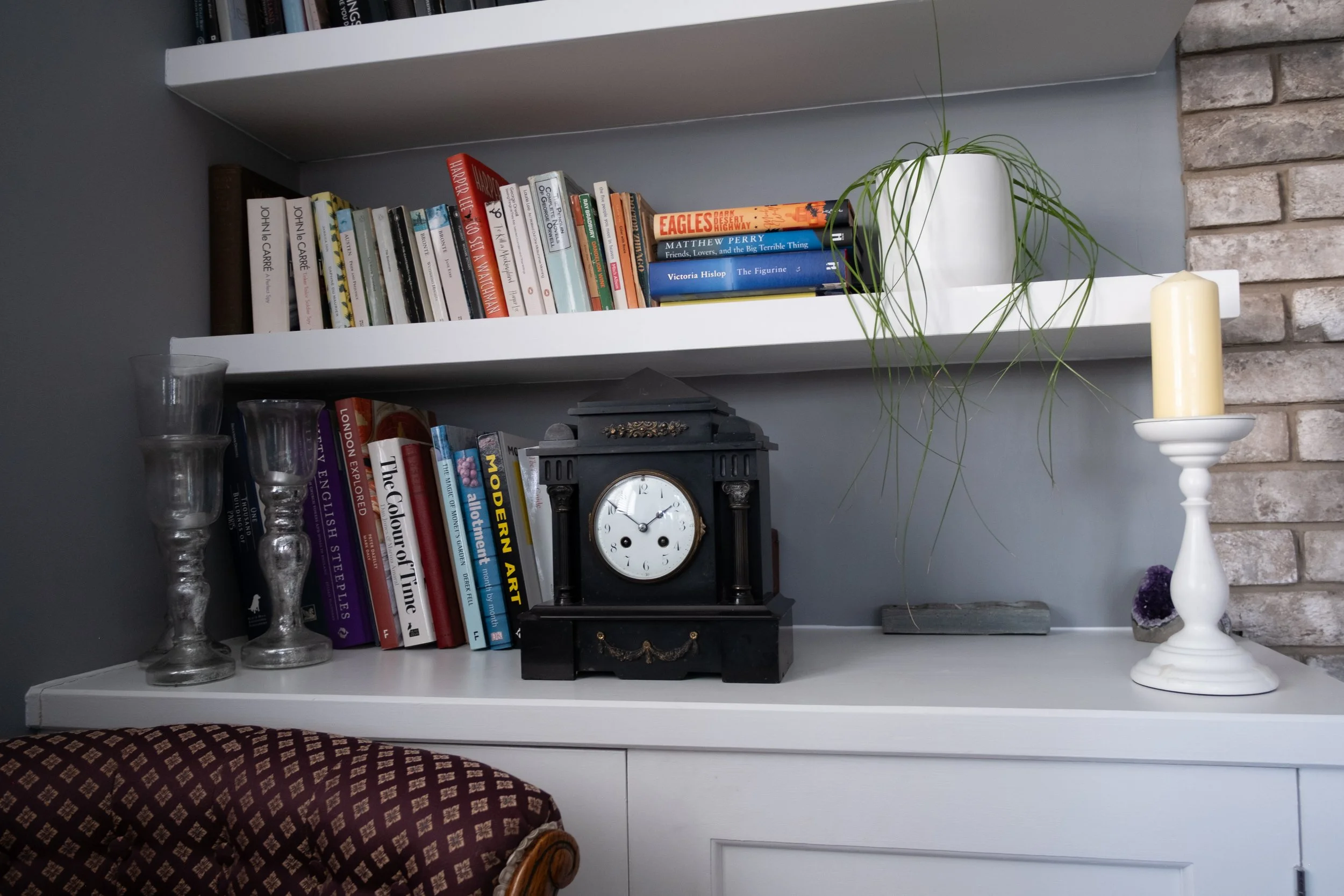 A white bookshelf with two shelves holding various books, a black antique clock, a potted plant, a white candlestick with a cream candle, and a geode. Part of a patterned armchair is visible in the foreground.