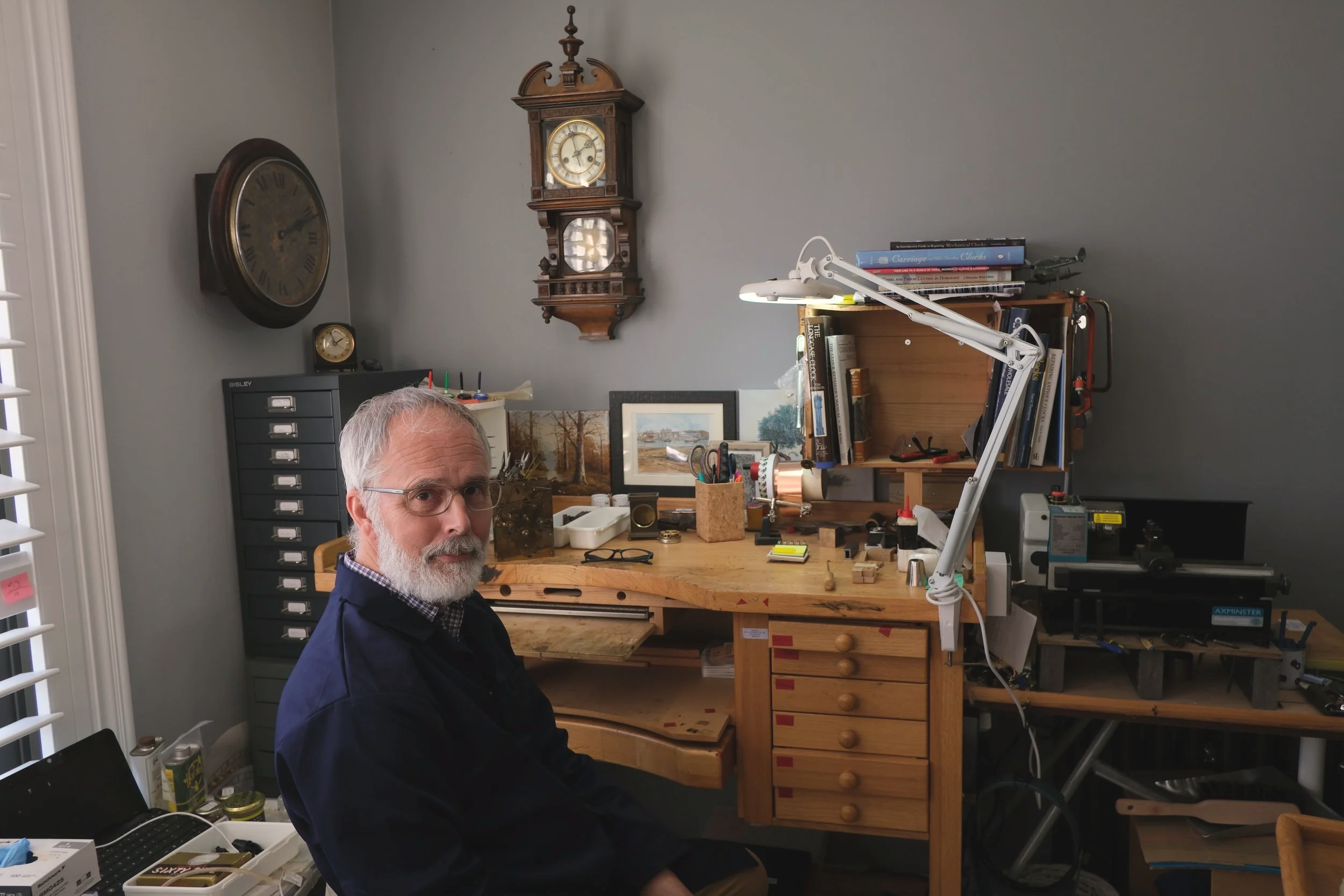 An older man with glasses and a beard is sitting at a wooden workbench in a room with gray walls. The desk has various tools, books, and artwork on it. Behind him, there are two clocks on the wall and a small dark cabinet with drawers.