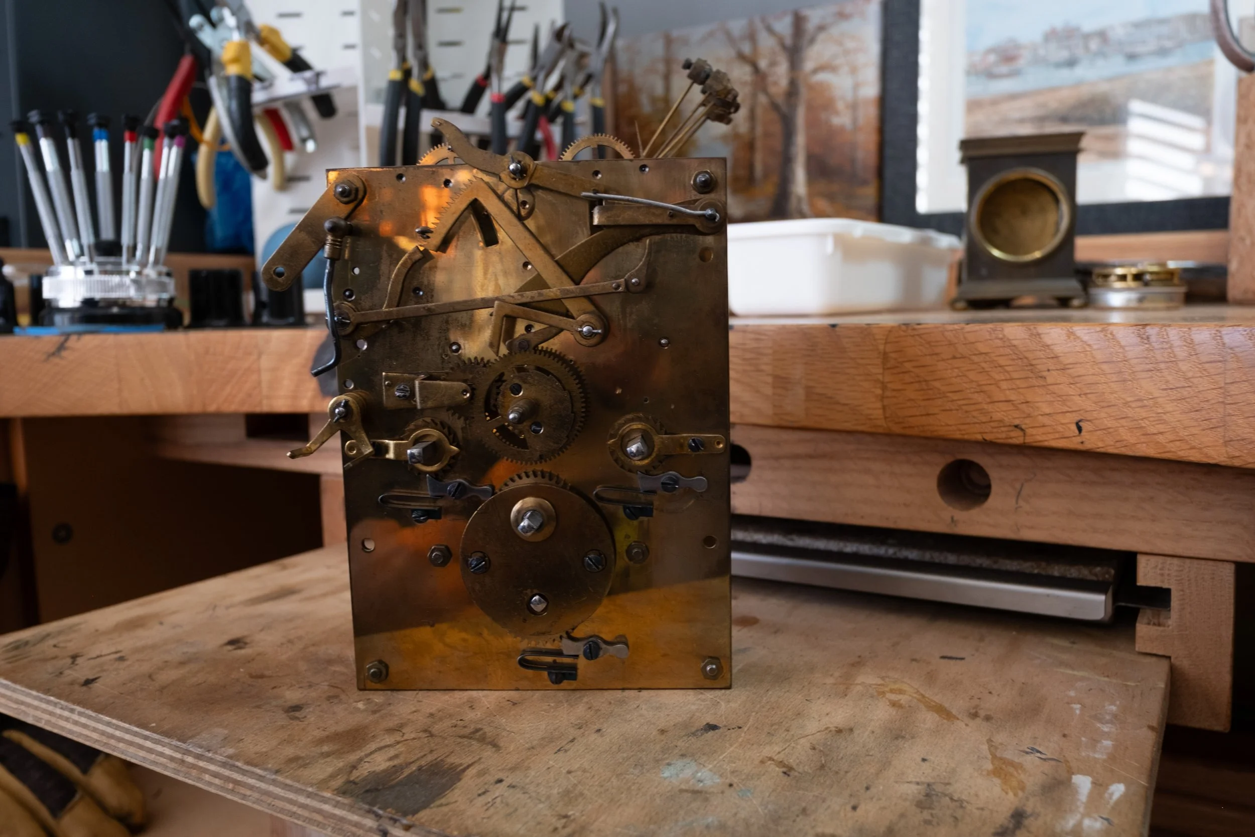 Close-up of a mechanical clock movement with gears and levers, placed on a wooden workbench in a workshop. In the background, tools and supplies are visible.