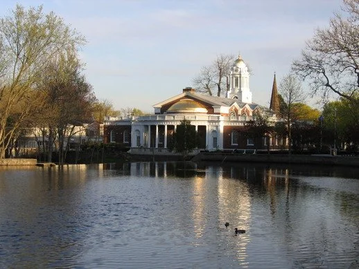 A historic building with a white dome and spire, surrounded by trees, reflected in a body of water, with ducks swimming nearby.
