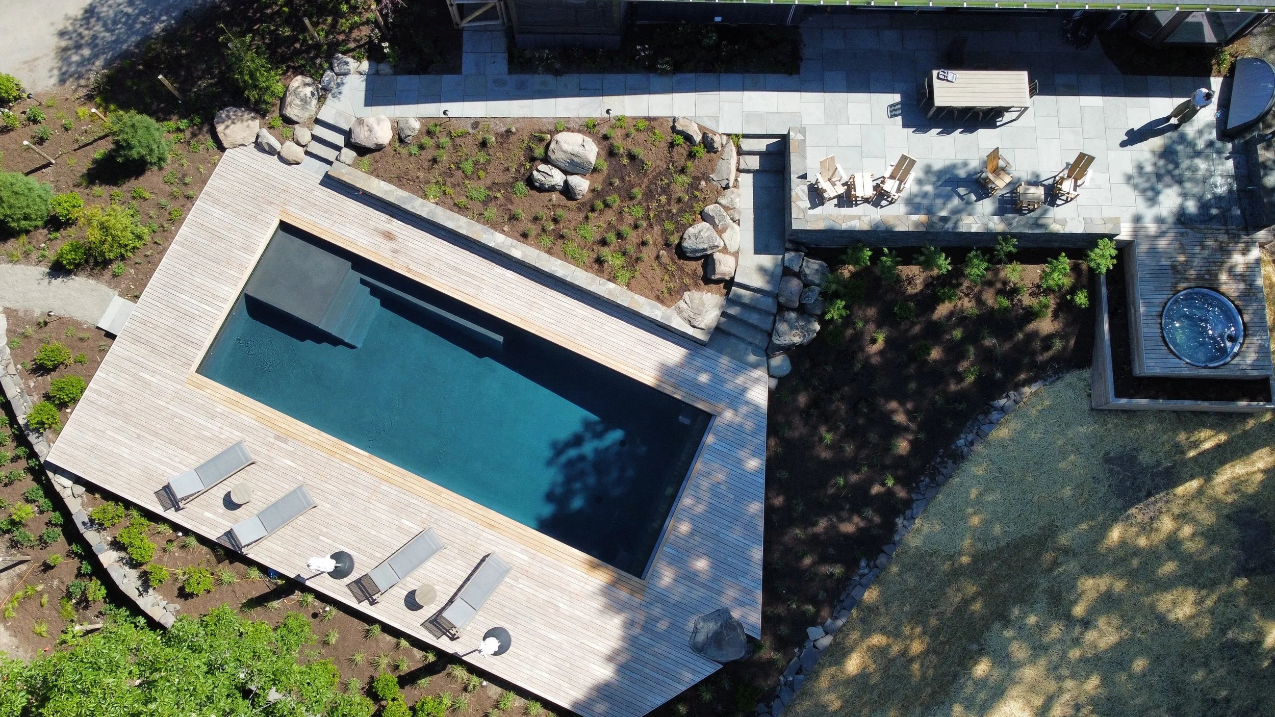 An outdoor swimming pool with a natural stone border and wooden decking, surrounded by trees and a forest in the background, under a clear sky.