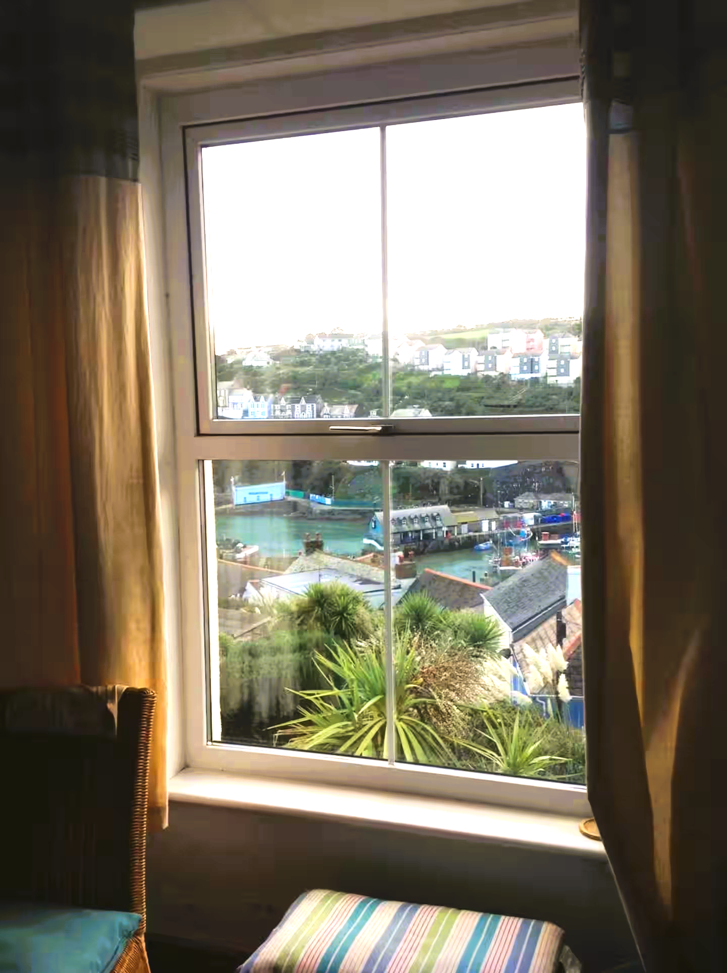View through a house window showing a neighborhood with houses, trees, a marina with boats, and a hill in the background. Inside, there's a chair with a striped cushion and curtains on either side of the window.