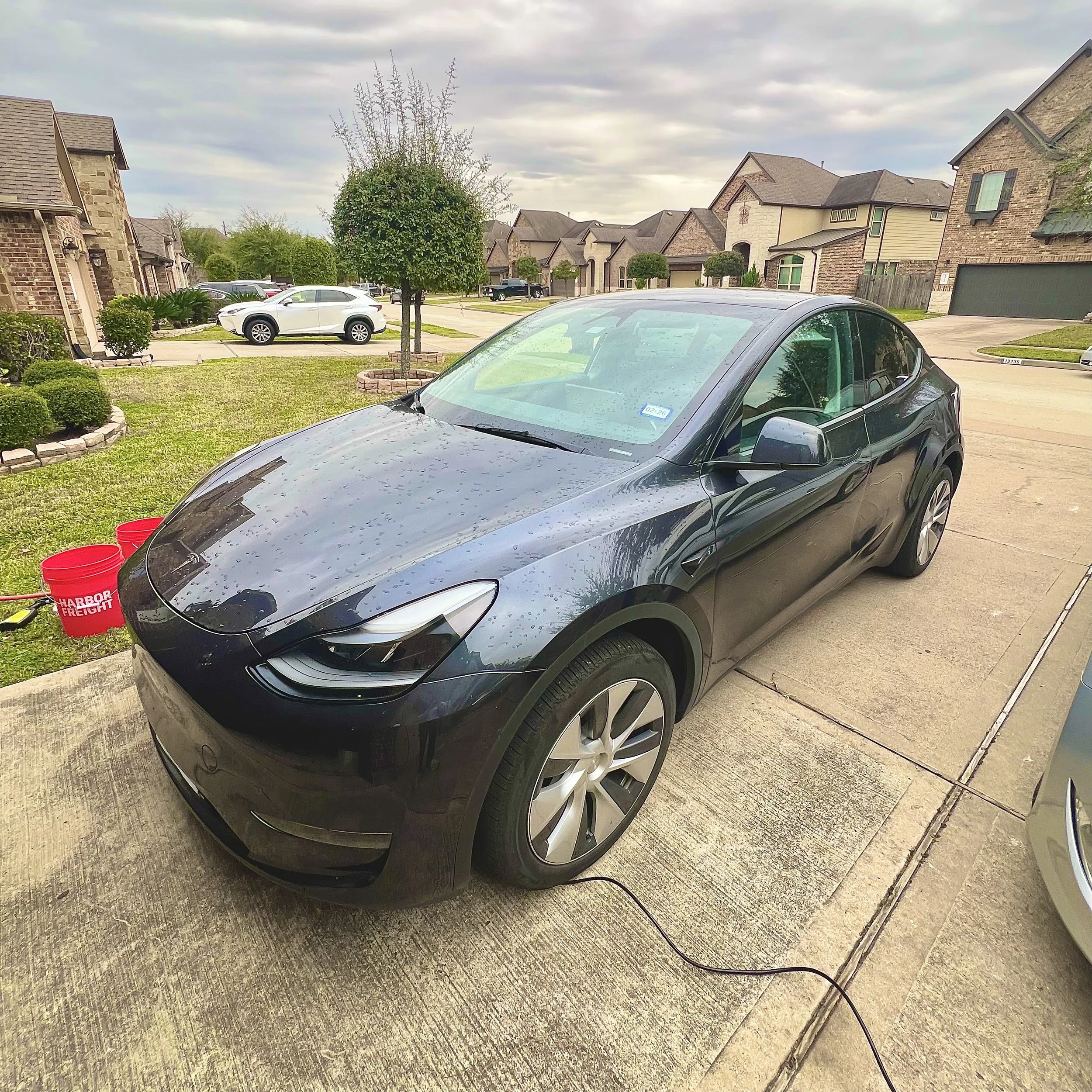 Black electric car parked in driveway with charging cord, in a suburban neighborhood with houses, trees, and other parked cars in the background, and red buckets nearby.