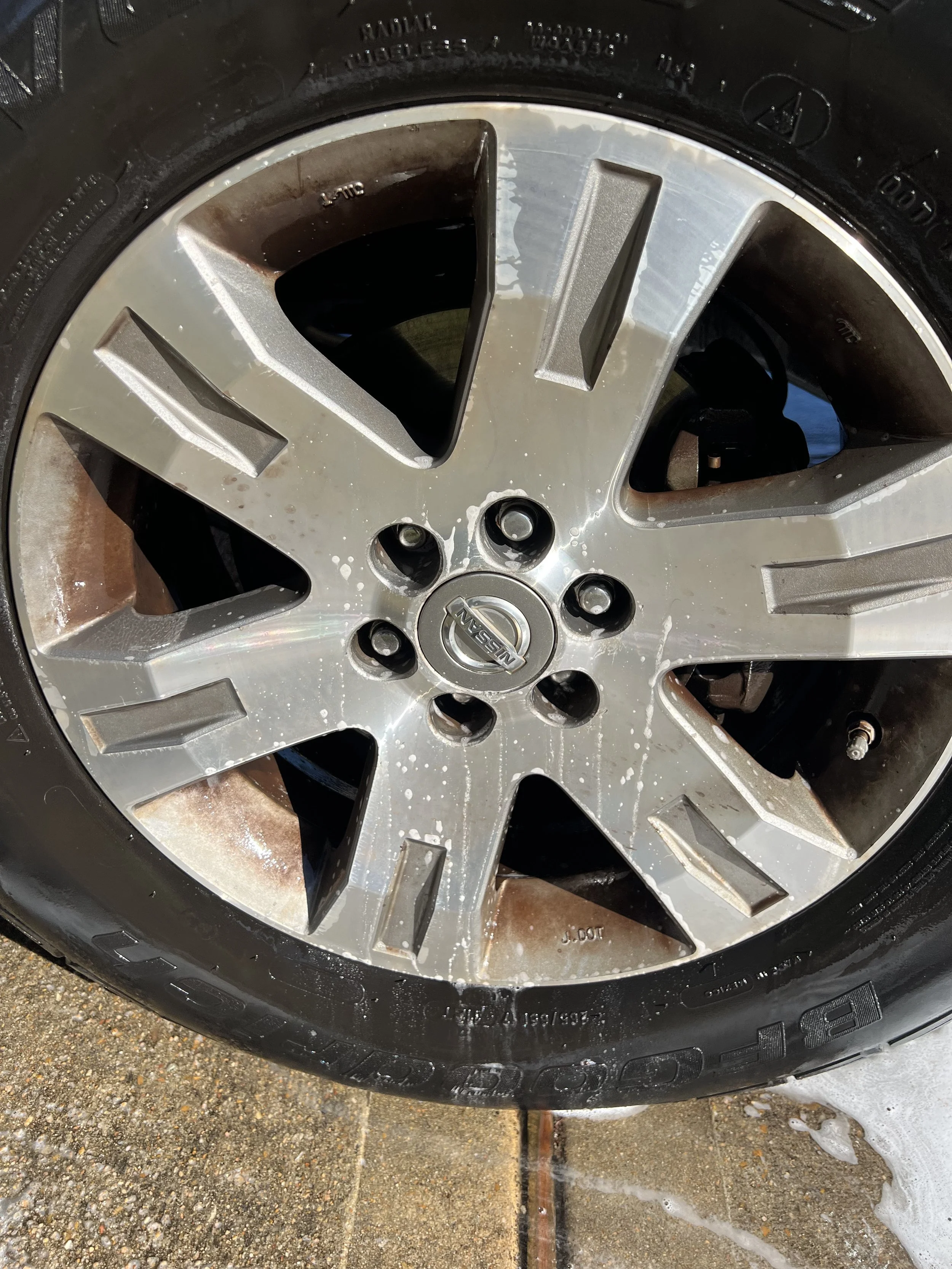 Close-up of a Nissan vehicle wheel with a silver alloy rim and black tire, showing soap water and dirt splatters on the rim, on a concrete surface.