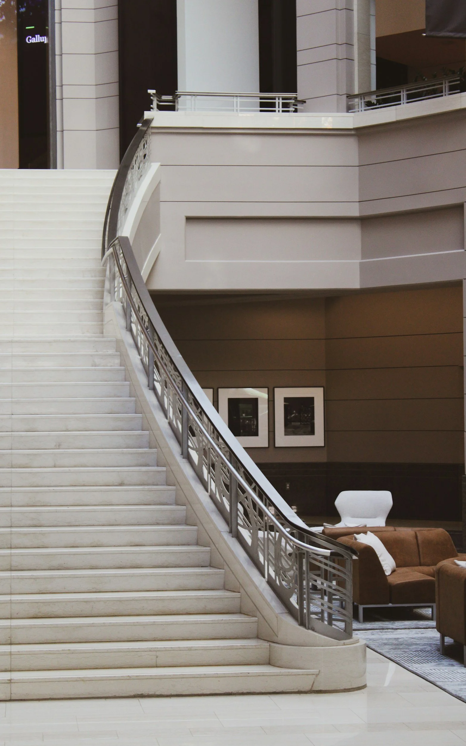 Marble staircase with metal railing inside a modern building, with seating and framed pictures on the wall in the background.