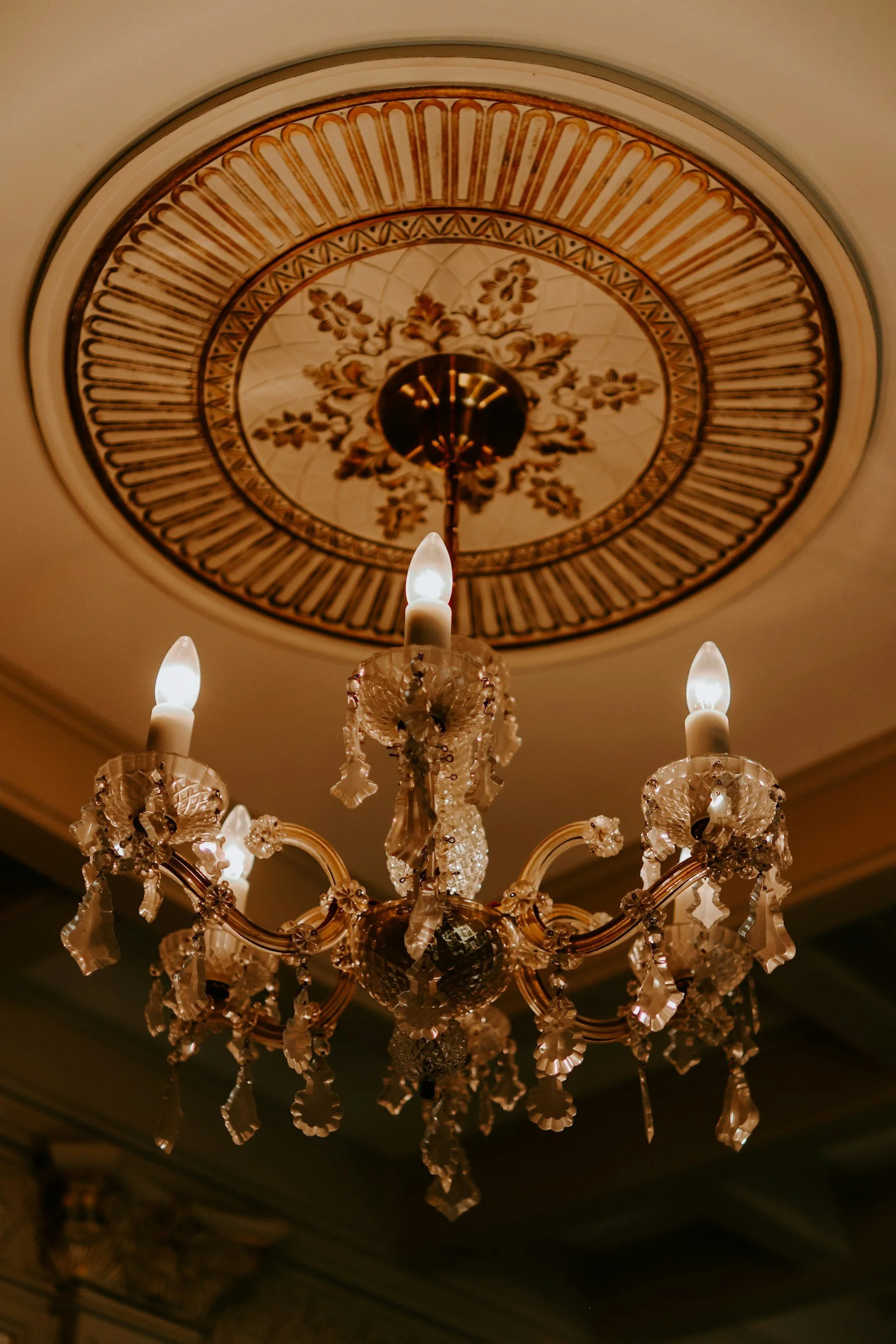 A decorative chandelier with lit candle-shaped light bulbs hanging from a ceiling with a vintage floral design in an ornate room.