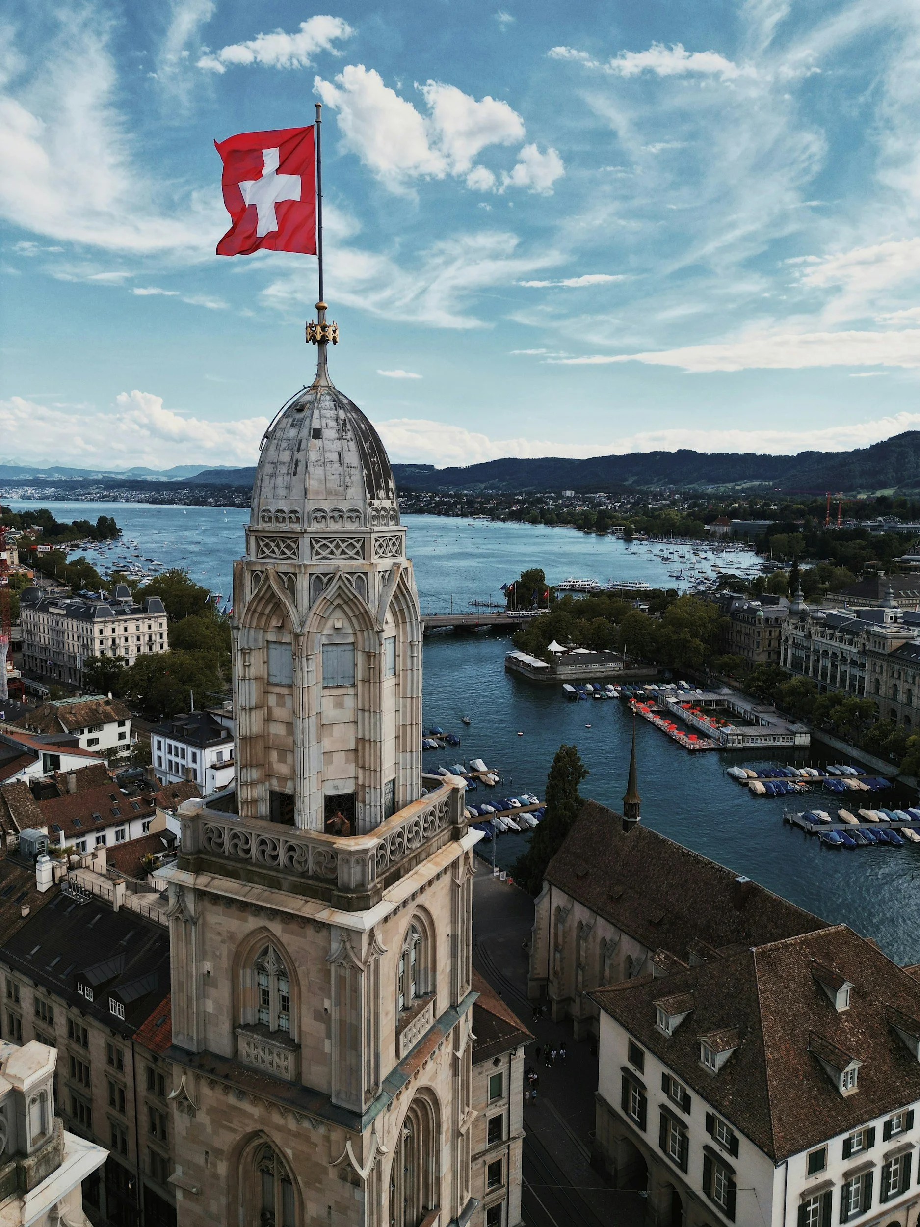 Aerial view of a historic building with a clock tower topped by a Swiss flag, overlooking a river and cityscape with boats and mountains in the background.