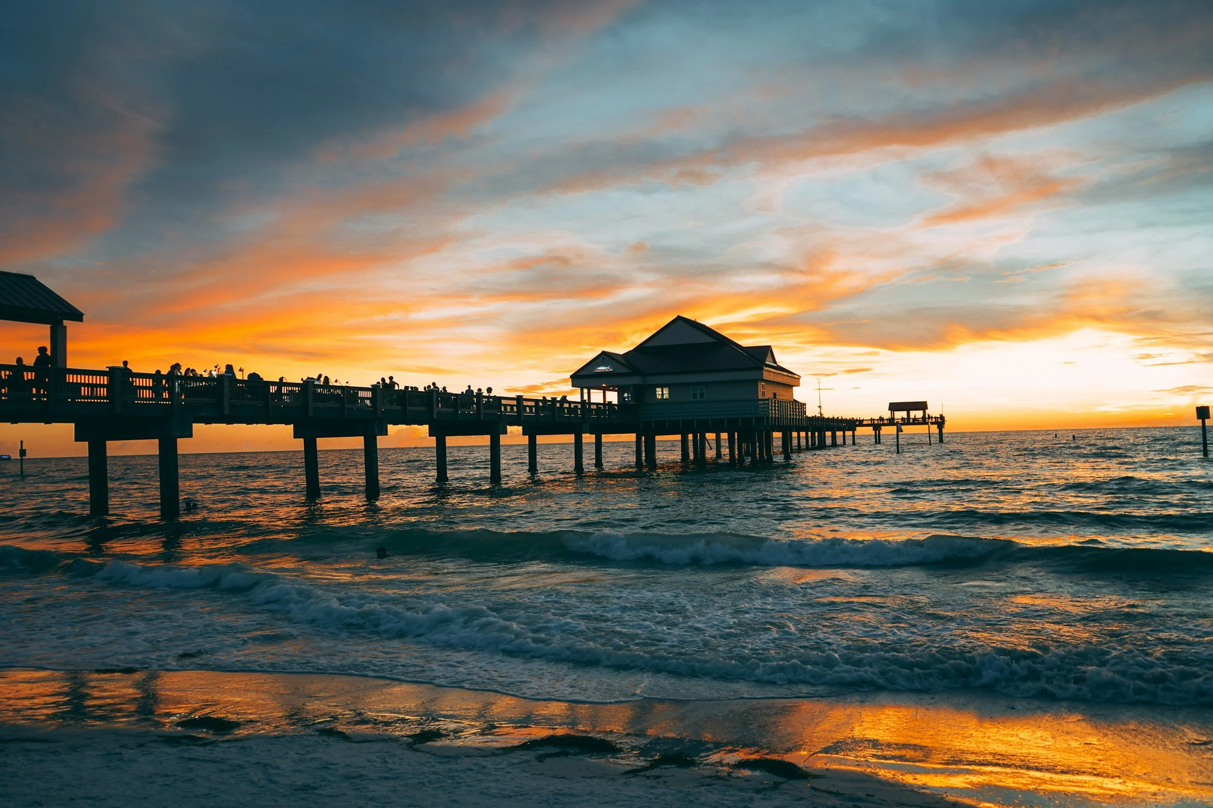 Sunset over the ocean with a pier extending into the water, a small building on the pier, and silhouettes of people watching the sunset.