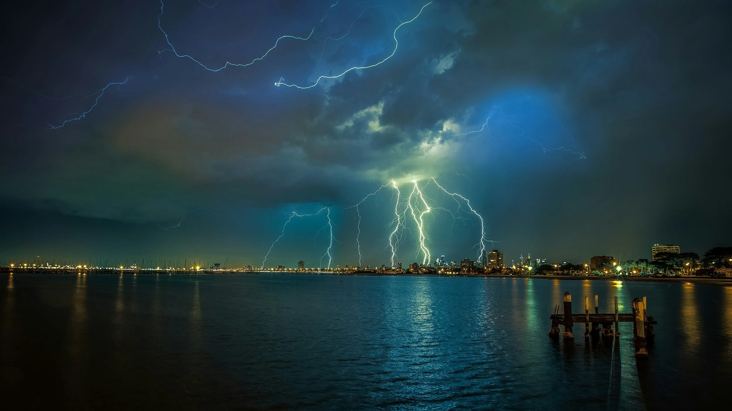 Nighttime cityscape with lightning strikes in the stormy sky over a body of water.