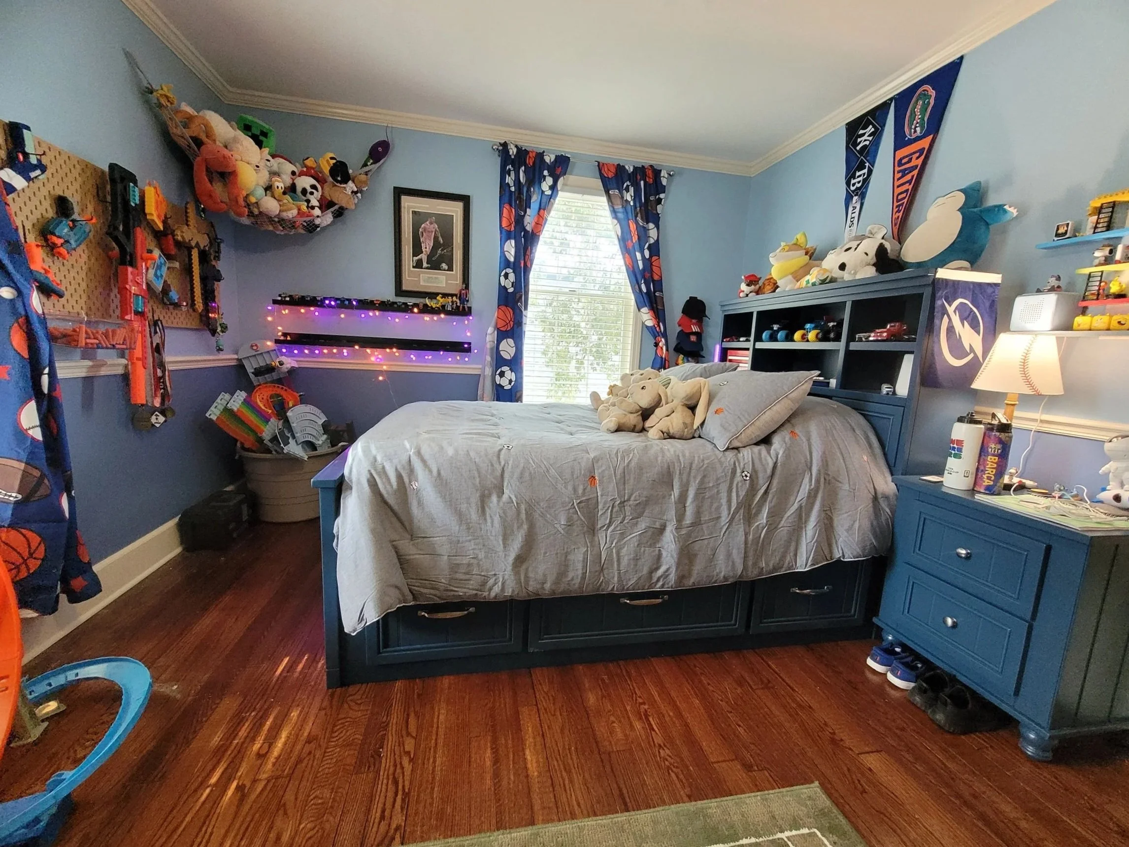 A blue bedroom with toys and football themed curtains, a bed with stuffed animals, a blue dresser, and a window with blinds.