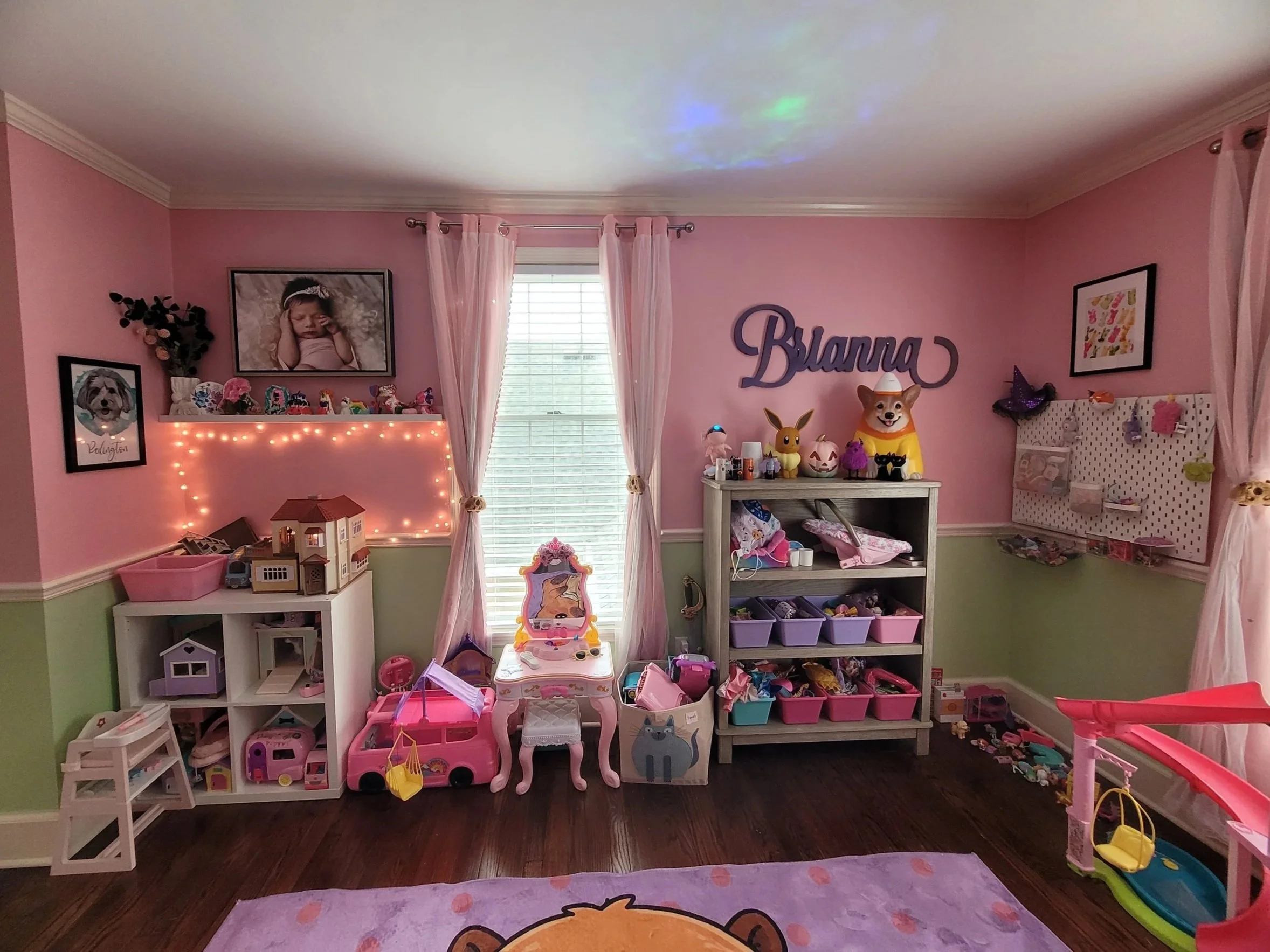 A pink-themed girl’s bedroom with stuffed animals, toys, and wall decorations, including framed photos and artwork, with curtains and a window in the background.
